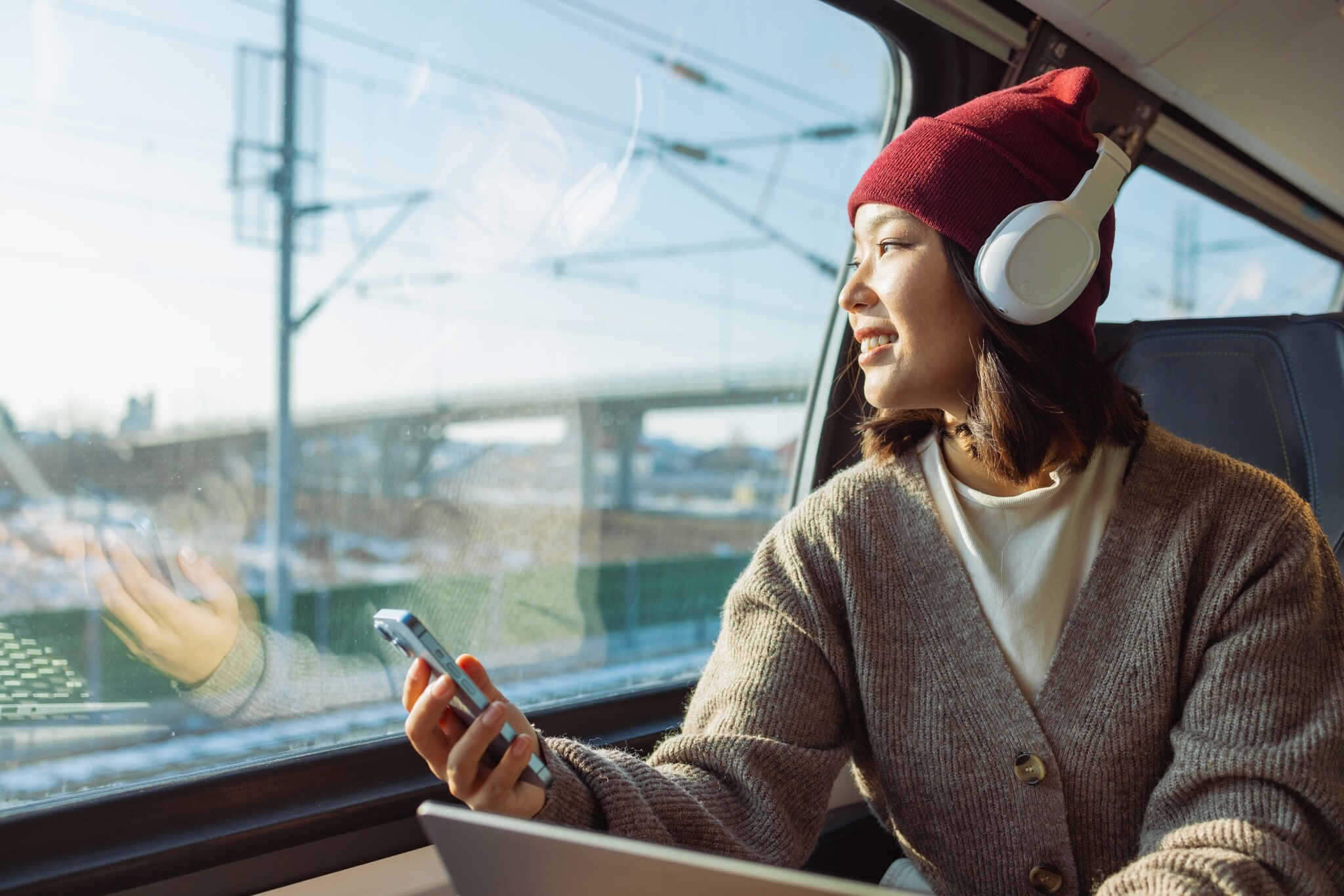 Eine Person mit Kopfhörer und Smartphone sitz in einem Zug. Sie trägt eine rote Mütze und einen grauen Pullover. Im Hintergrund sind Züge und Schienen zu sehen. Eine Person mit Kopfhörer und Smartphone sitz in einem Zug. Sie trägt eine rote Mütze und einen grauen Pullover. Im Hintergrund sind Züge und Schienen zu sehen.
