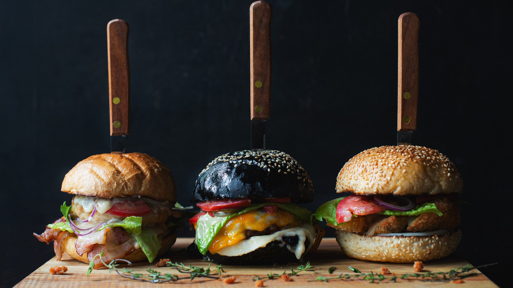 Drei verschiedene Burger mit Messern im Brötchen auf einem Holzbrett, darunter Salat, Tomate und Käse sichtbar