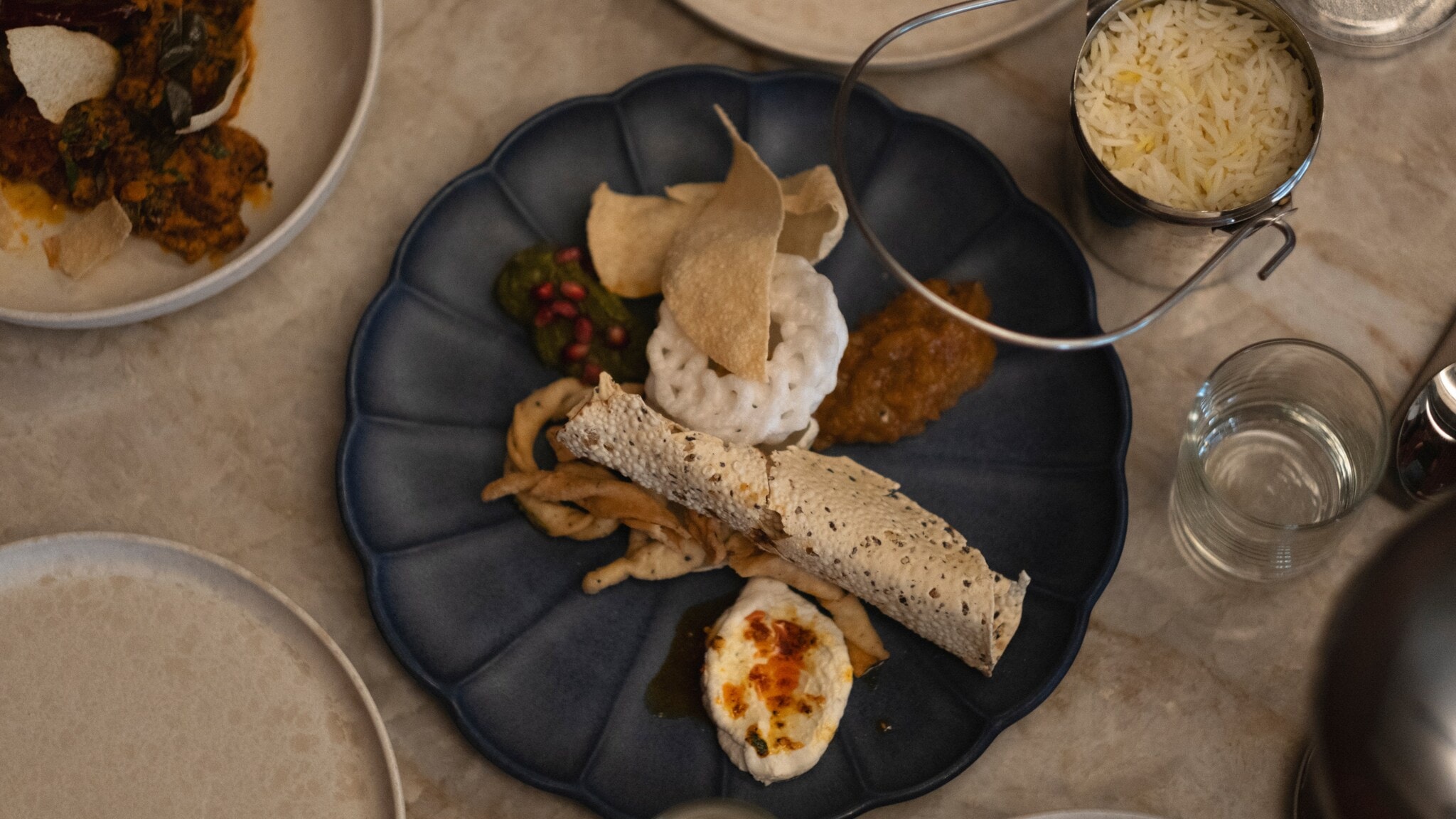 Ein blauer Teller mit verschiedenen indischen Gerichten: Reis in einer Schüssel, Papadam, Chutney, frittiertes Gemüse und ein gerolltes Fladenbrot.