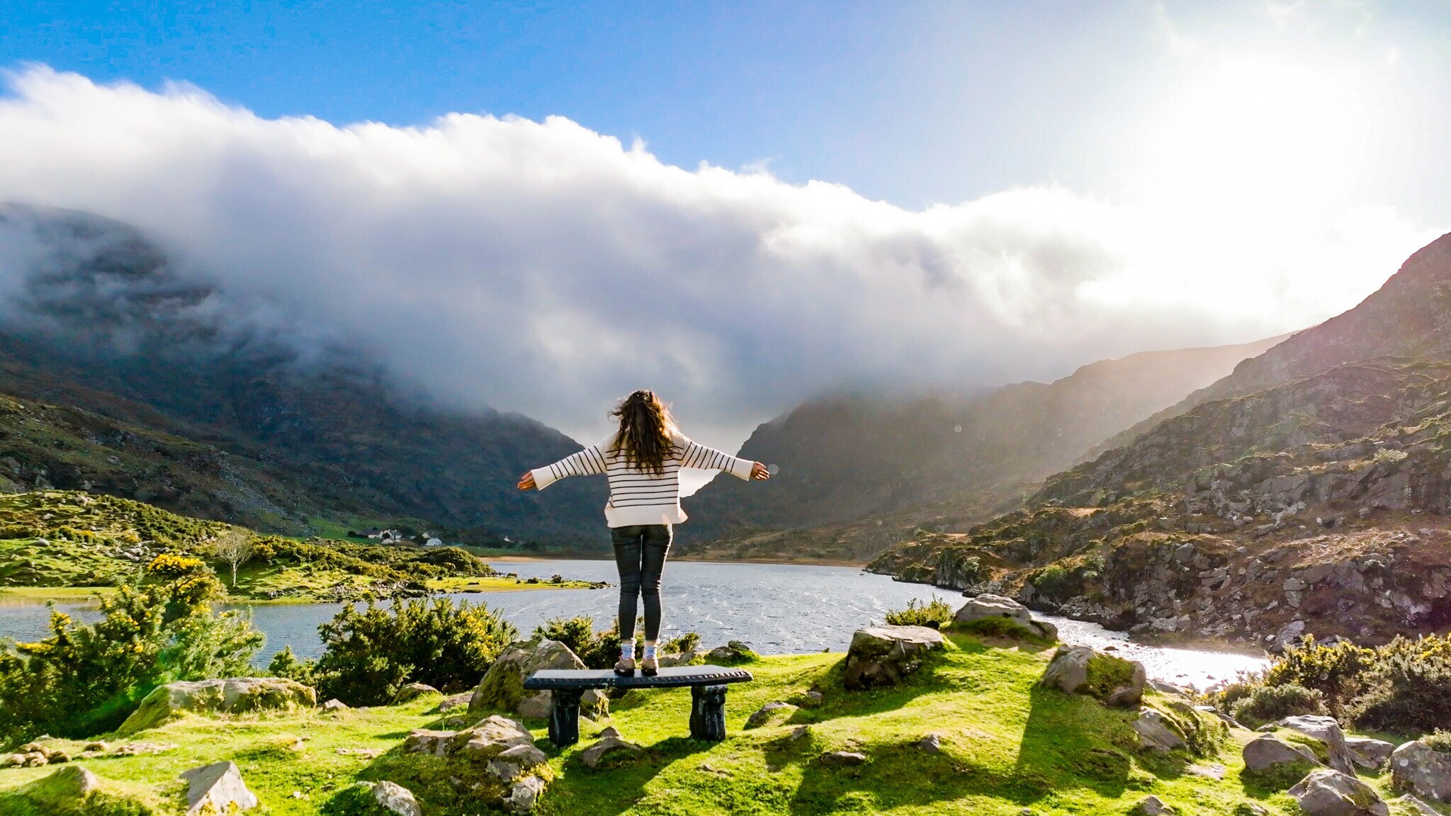 Eine Person mit langen, offenen Haaren steht auf einem Stein und breitet die Arme aus. Sie trägt ein gestreiftes Oberteil und blickt auf einen See, umgeben von Bergen und Wolken. Eine Person mit langen, offenen Haaren steht auf einem Stein und breitet die Arme aus. Sie trägt ein gestreiftes Oberteil und blickt auf einen See, umgeben von Bergen und Wolken.