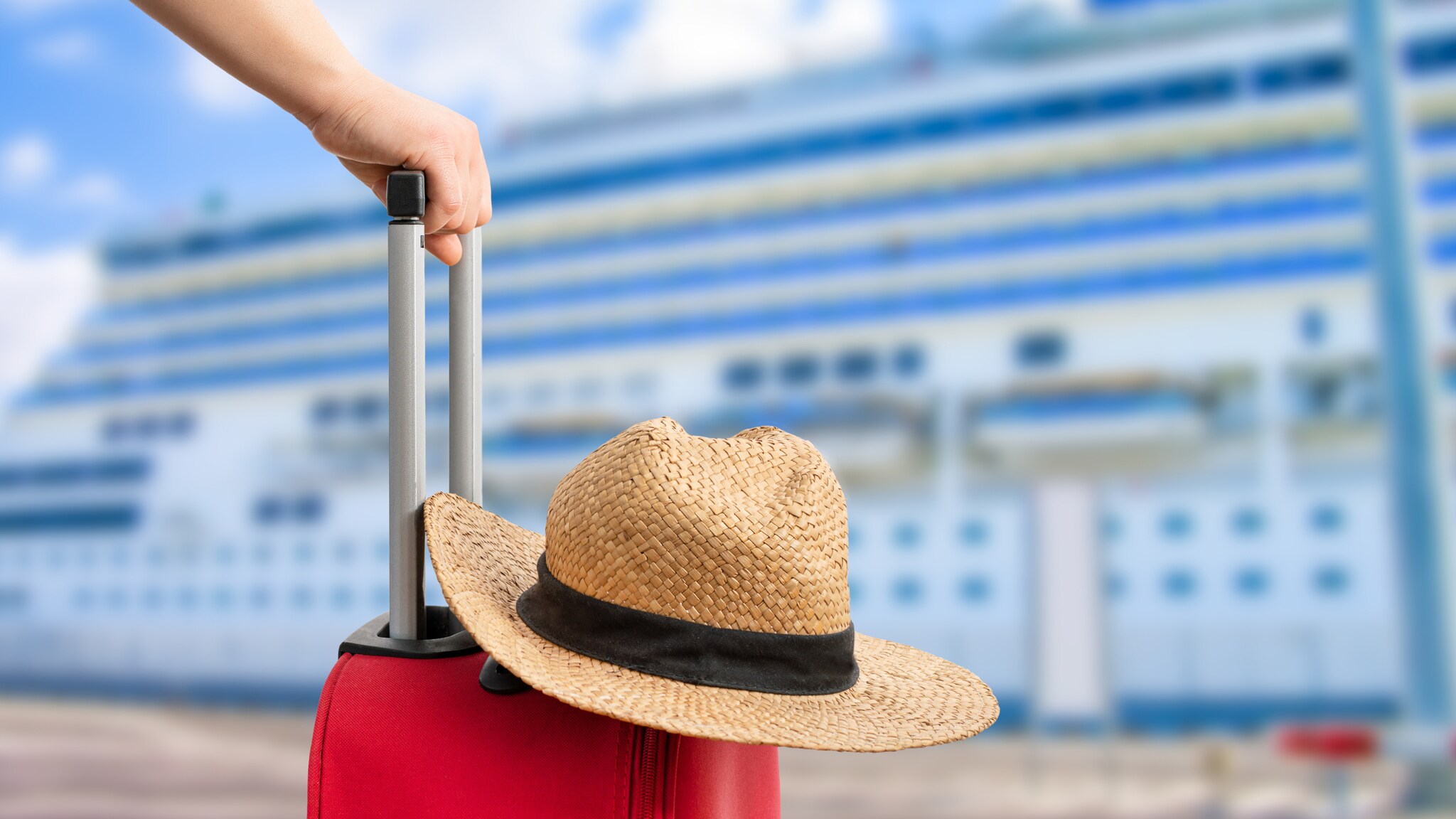 Hand hält Griff eines roten Rollkoffers, auf dem ein Strohhut mit schwarzem Band liegt, im Hintergrund ein Kreuzfahrtschiff.