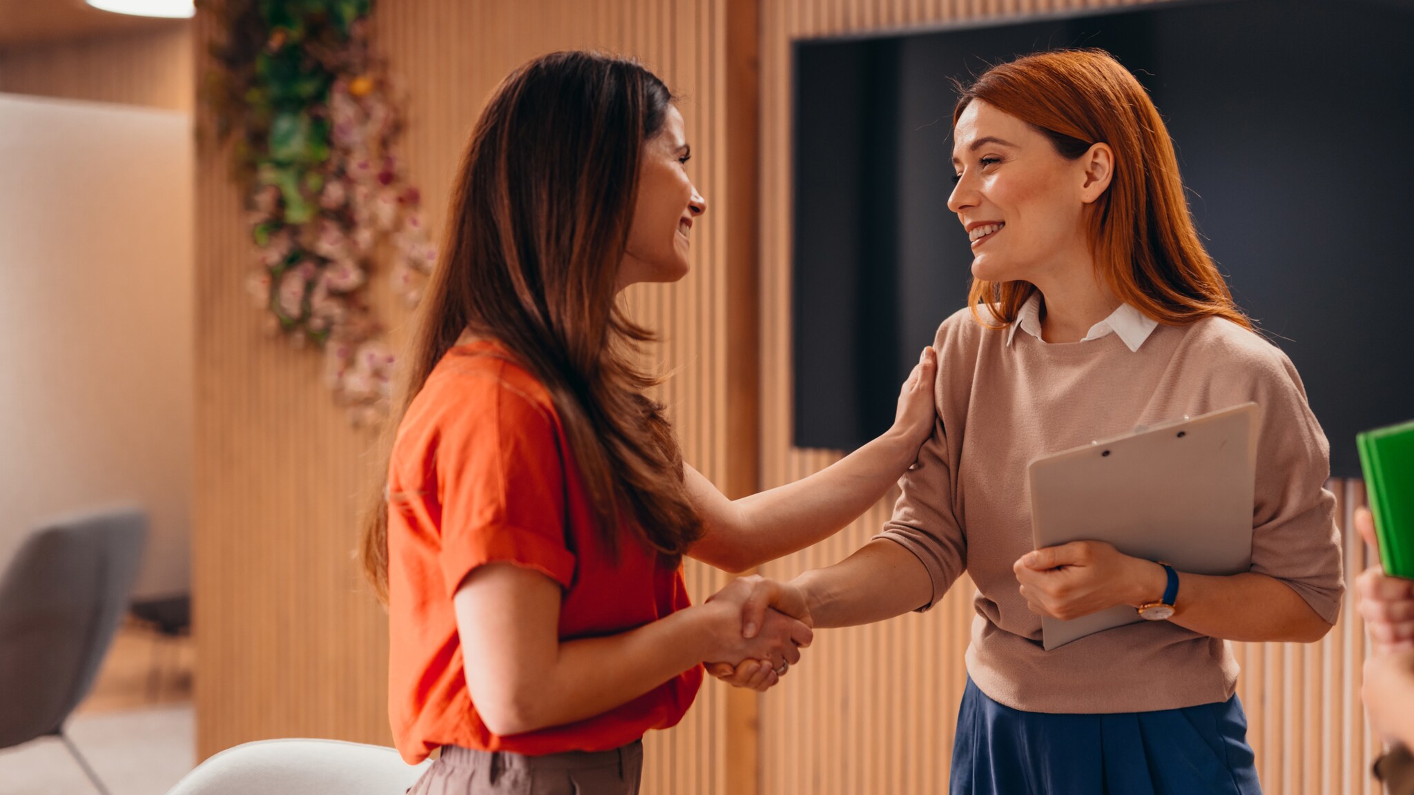 Zwei Frauen geben sich die Hand und lächeln sich an. Eine Frau legt die Hand auf den Arm der anderen Frau.