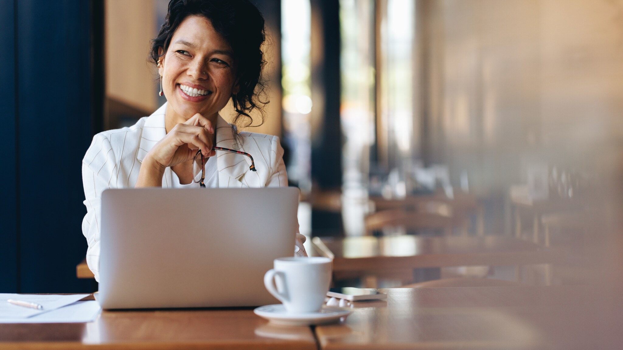 Eine Frau sitzt zufrieden lächelnd an einem Laptop an einem Tisch mit Kaffeetasse in einem Café. Eine Frau sitzt zufrieden lächelnd an einem Laptop an einem Tisch mit Kaffeetasse in einem Café.