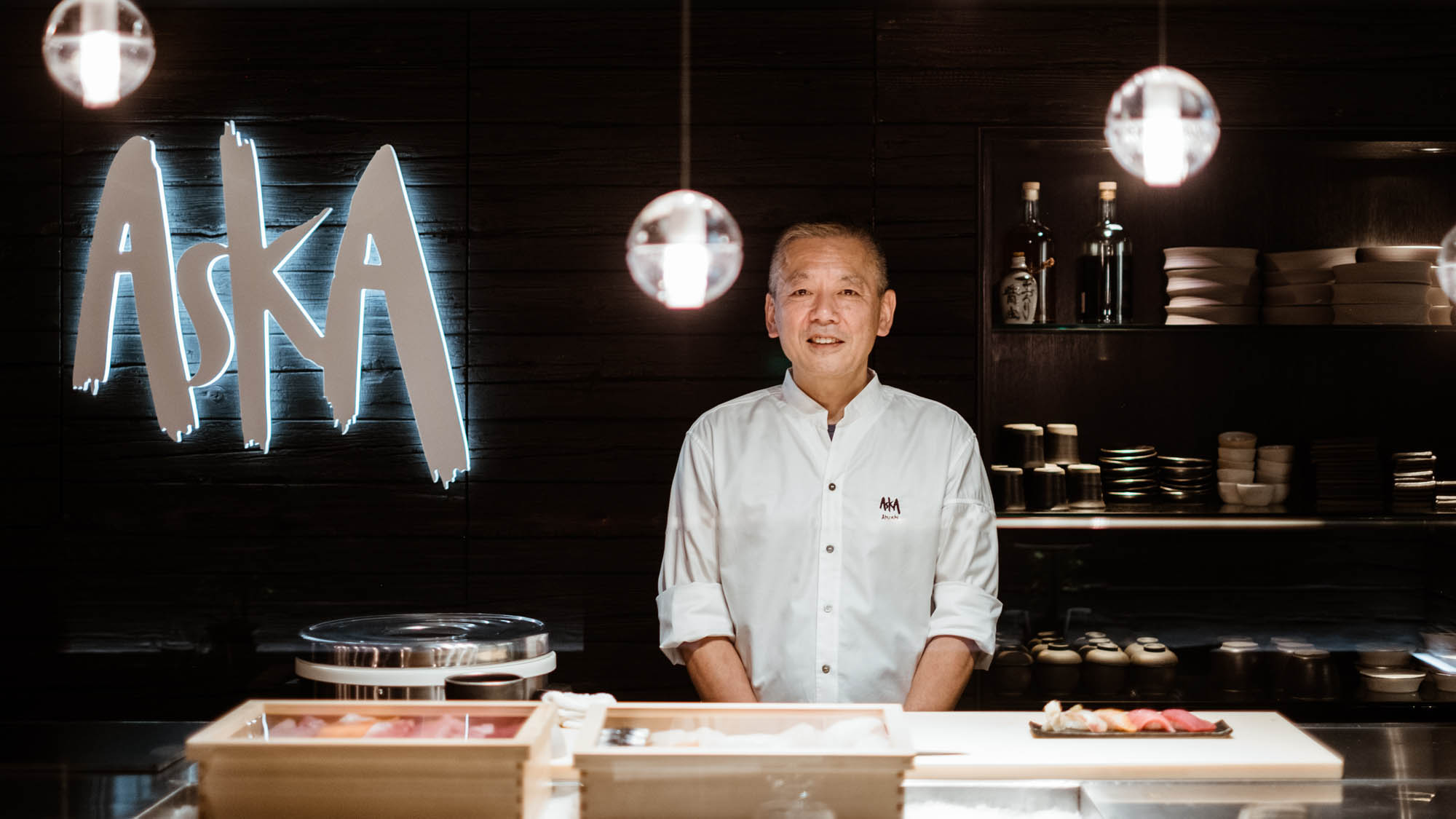 Ein Küchenchef steht hinter einer Sushibar in einem Restaurant mit einem Wandlogo Aska.