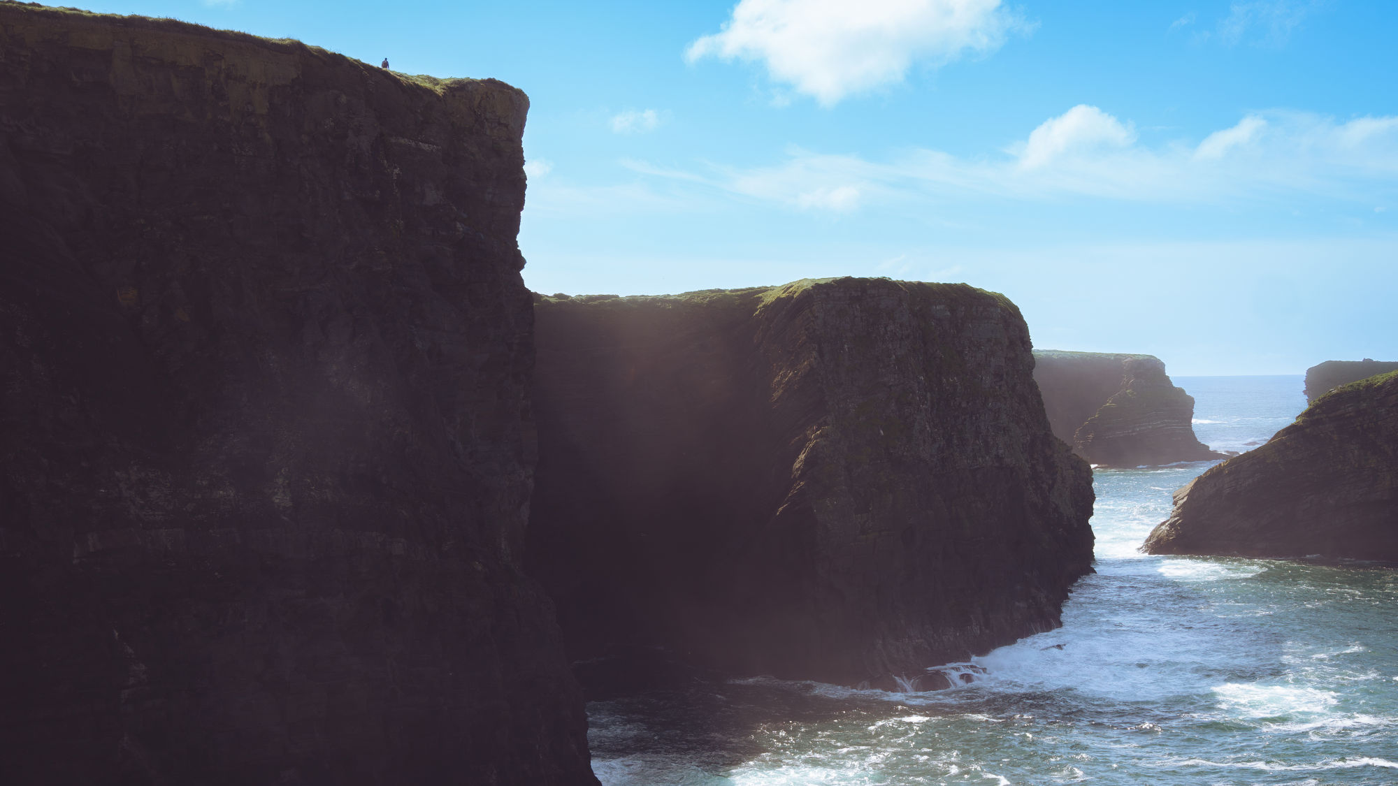 Hohe Klippen an der Küste von Kilkee mit Wellen, die gegen die Felsen schlagen, unter blauem Himmel. Hohe Klippen an der Küste von Kilkee mit Wellen, die gegen die Felsen schlagen, unter blauem Himmel.