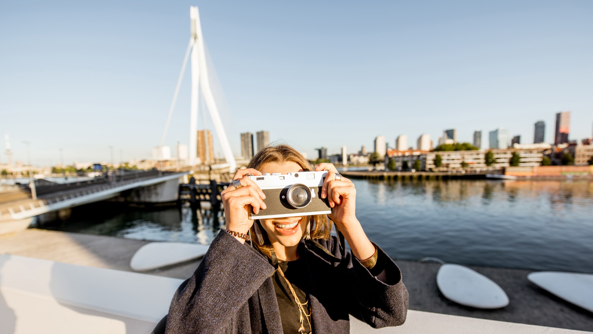 erson mit dunkelgrauem Mantel hält eine weiße Sofortbildkamera vor das Gesicht, im Hintergrund die Skyline von Rotterdam und eine Brücke über Wasser.