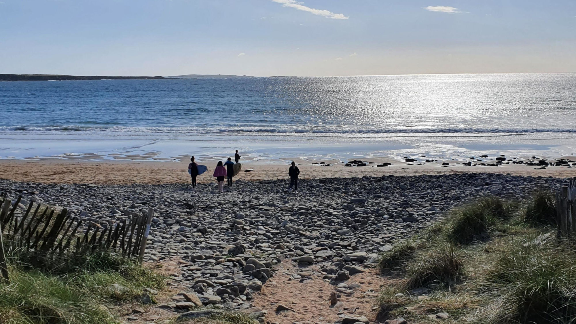 Blick auf den Strand von Spanish Point Beach mit mehreren Personen, die mit Surfbrett zum Wasser gehen, felsigem Ufer und reflektierender Meeresoberfläche bei klarem Himmel. Blick auf den Strand von Spanish Point Beach mit mehreren Personen, die mit Surfbrett zum Wasser gehen, felsigem Ufer und reflektierender Meeresoberfläche bei klarem Himmel.