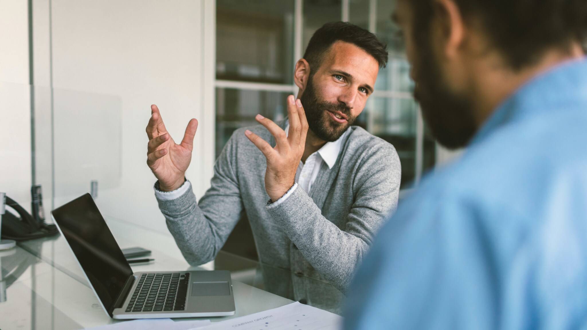 Ein Mann gestikuliert mit den Händen während eines Gesprächs mit einer Person an einem Tisch mit Laptop.
