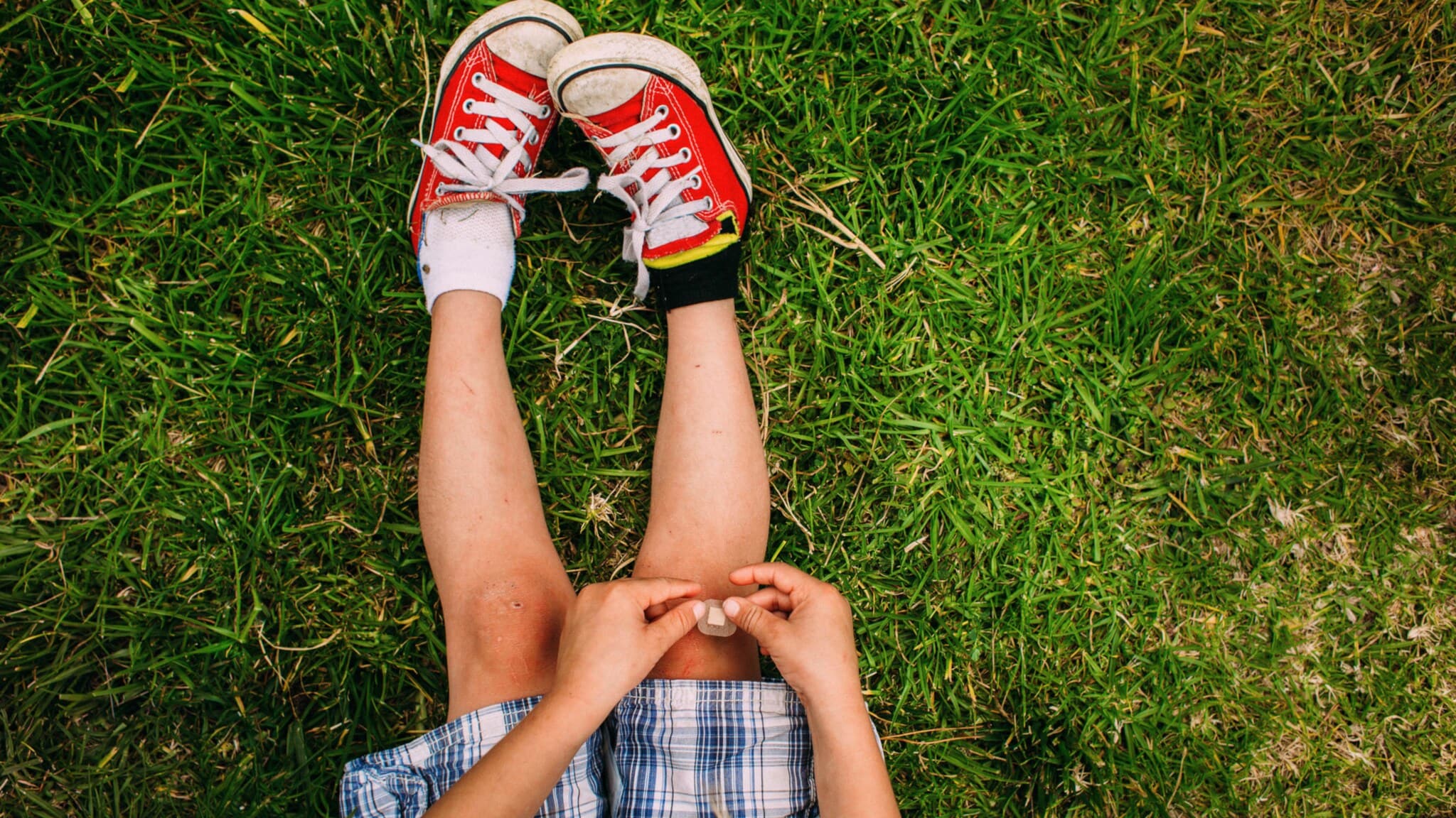Kind sitzt auf Gras, trägt rot-weiße Turnschuhe und blau karierten Rock, hält Pflaster an Knie. Kind sitzt auf Gras, trägt rot-weiße Turnschuhe und blau karierten Rock, hält Pflaster an Knie.