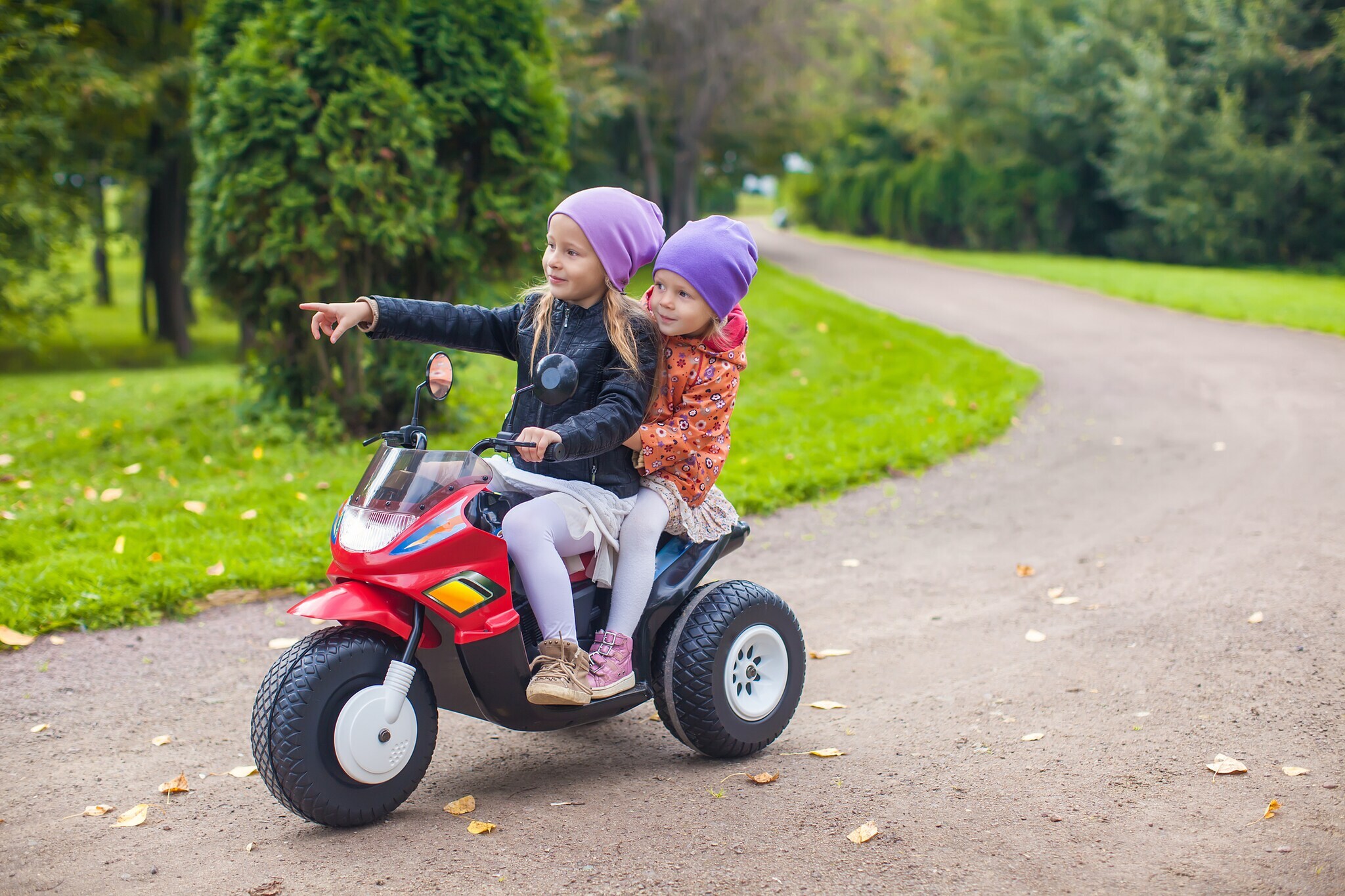 Zwei Mädchen fahren auf einem elektrischen Dreirad auf einem Sandweg.