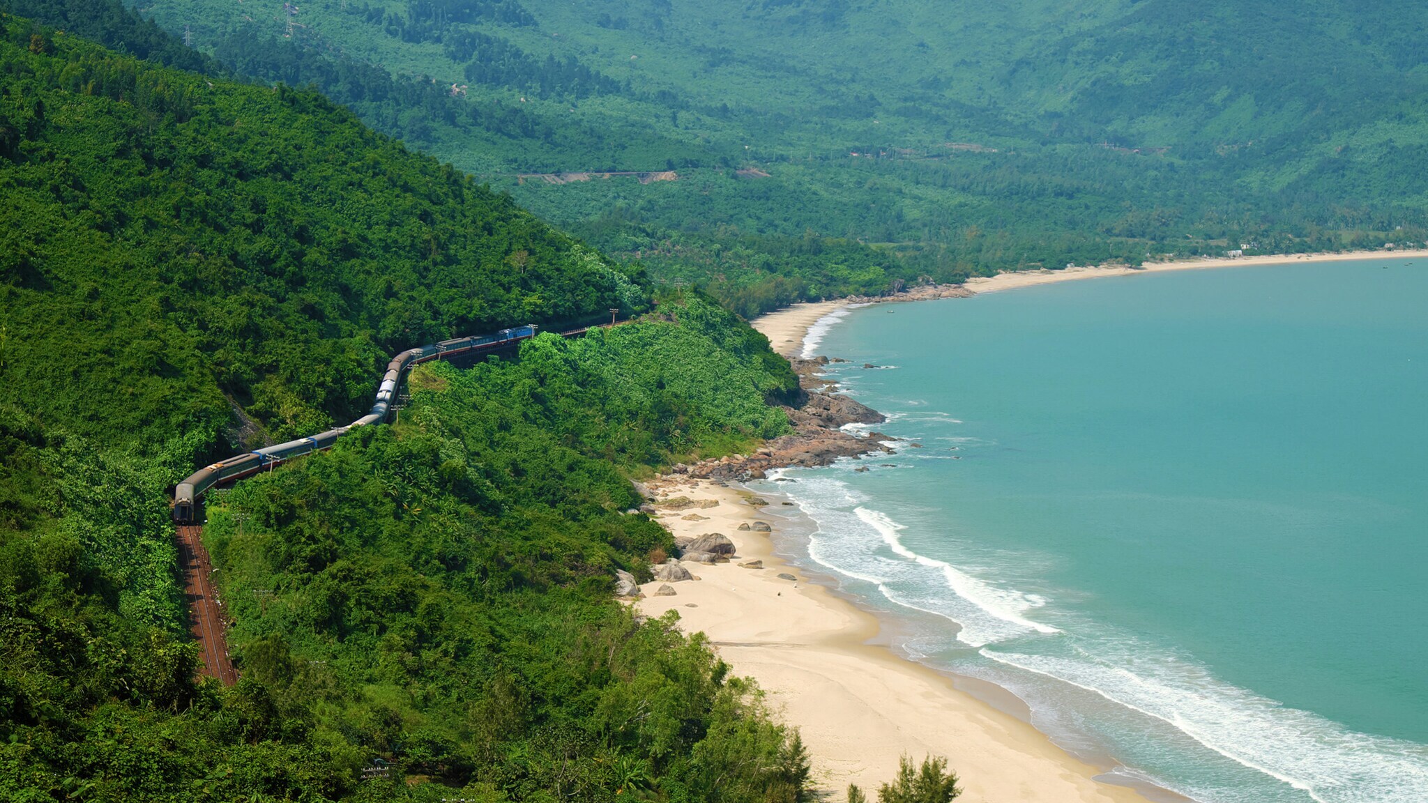 Ein Zug fährt entlang einer kurvenreichen Küstenlinie, umgeben von grünen Hügeln und einem Sandstrand. Das Wasser ist blau und die Wellen sind sichtbar.