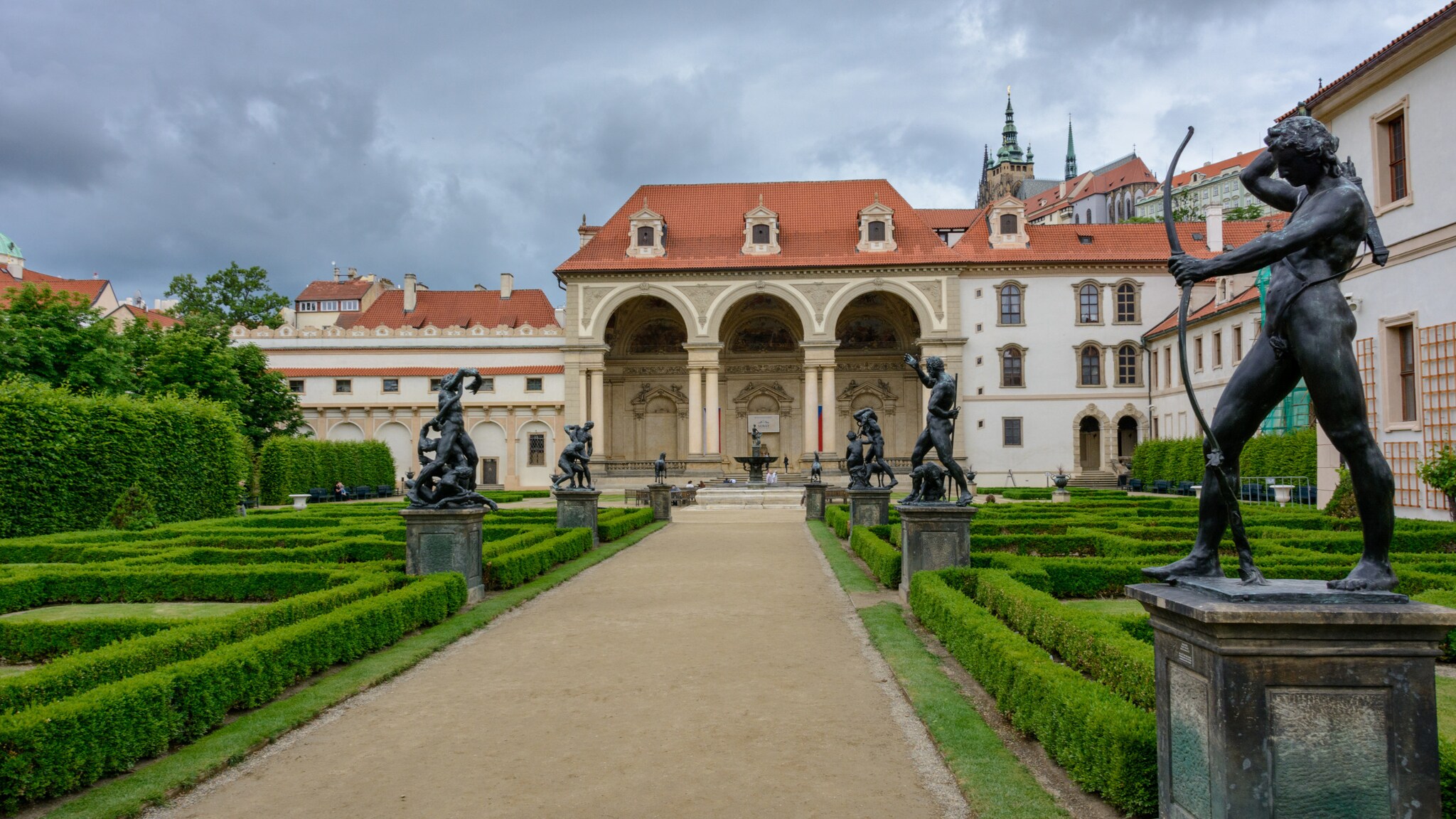 Barockgarten mit symmetrischen Hecken und mehreren Statuen vor einem historischen Gebäude mit Arkaden und rotem Ziegeldach unter bewölktem Himmel Barockgarten mit symmetrischen Hecken und mehreren Statuen vor einem historischen Gebäude mit Arkaden und rotem Ziegeldach unter bewölktem Himmel