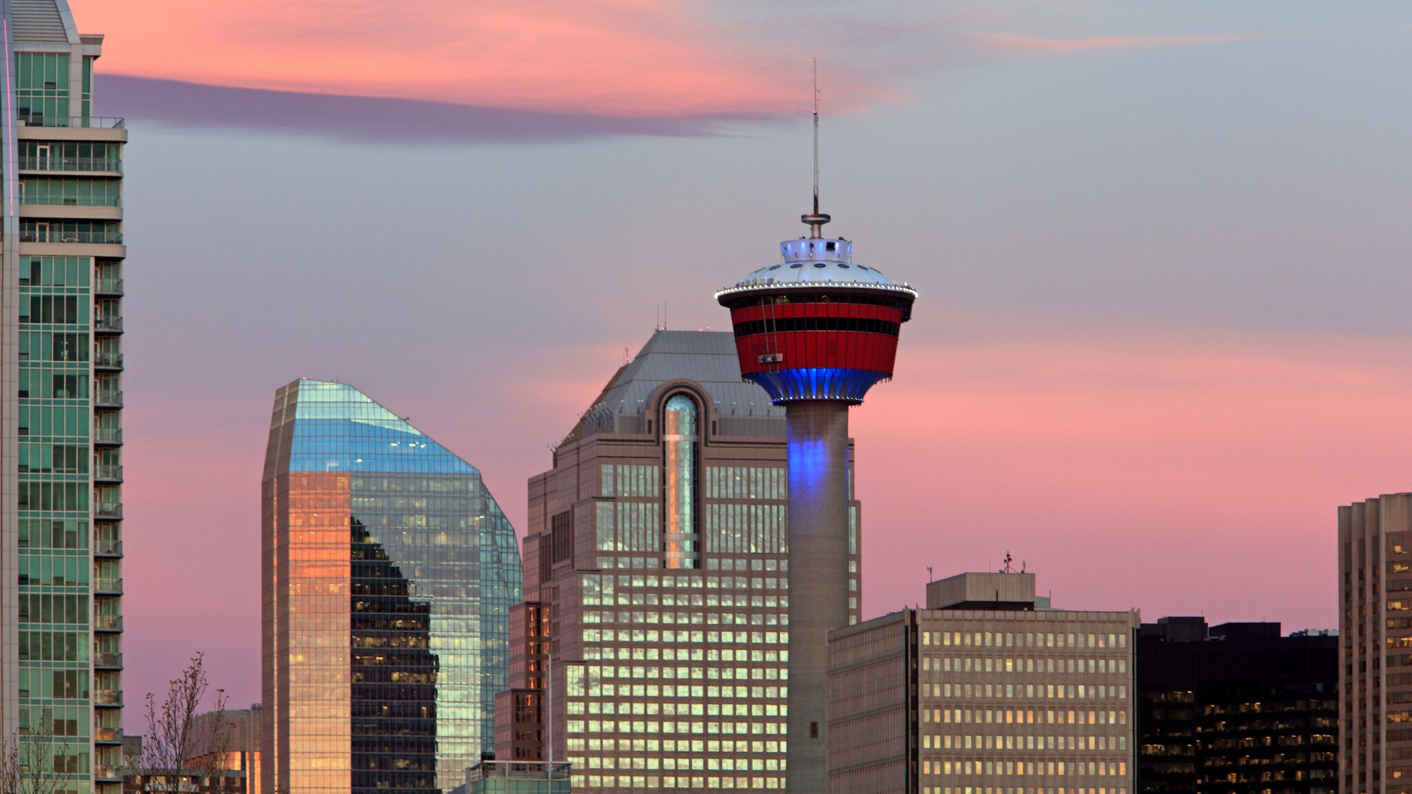Oberer Teil eines Fernsehturms mit rotem Turmkopf vor Hochhäusern mit glänzenden Fassaden im Abendlicht vor Himmel mit rosa gefärbten Wolken.