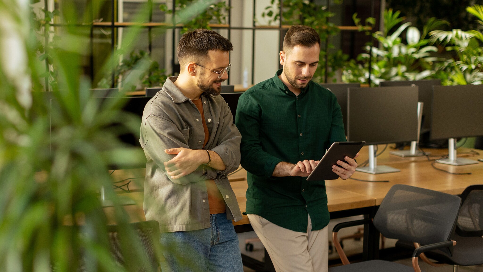 Zwei junge Männer lehnen an einem Schreibtisch mit vielen Arbeitsplätzen und schauen gemeinsam auf ein Tablet in einem modernen Großraumbüro. Zwei junge Männer lehnen an einem Schreibtisch mit vielen Arbeitsplätzen und schauen gemeinsam auf ein Tablet in einem modernen Großraumbüro.