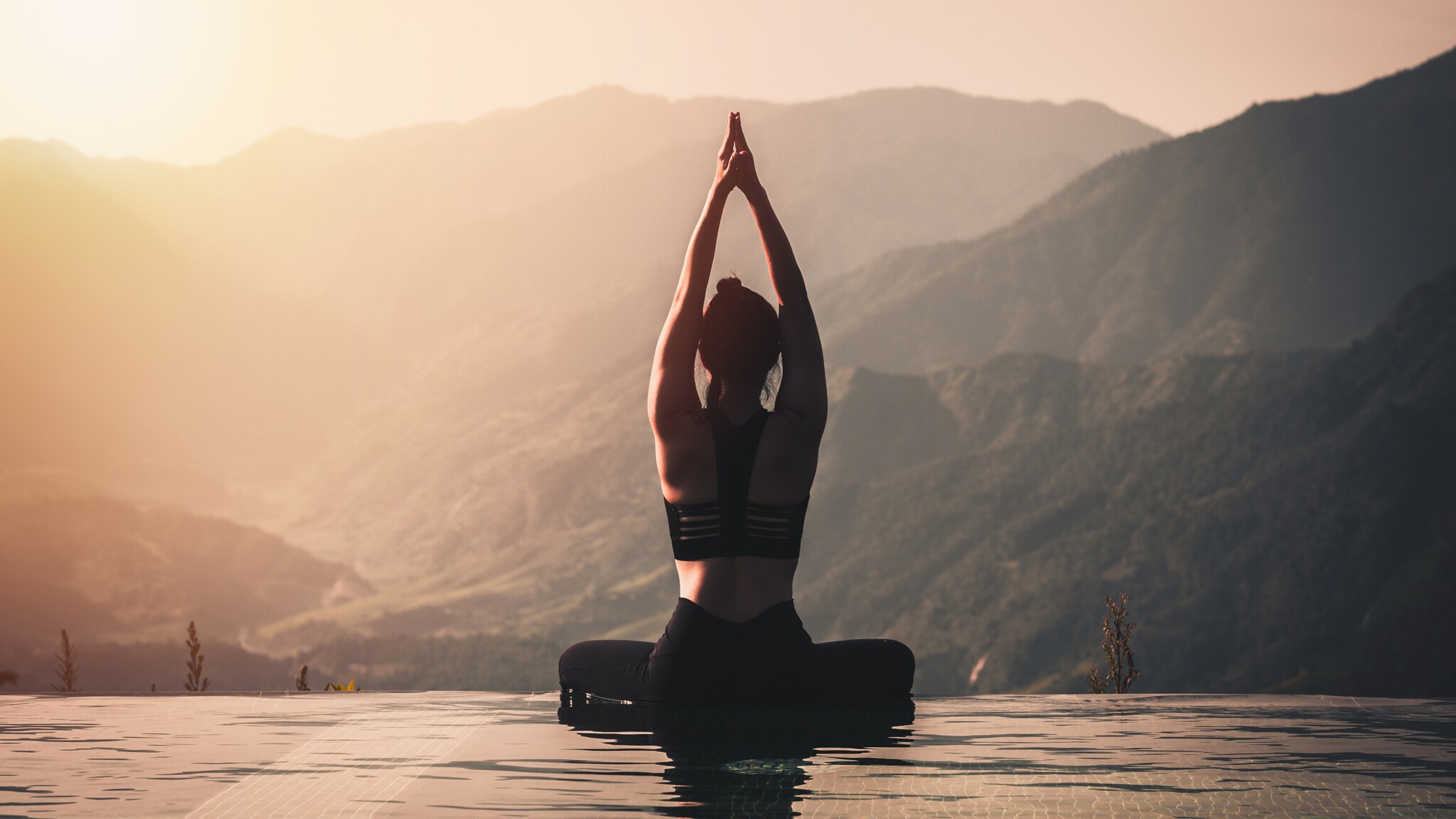 Eine Person in schwarzer Sportbekleidung sitzt im Lotussitz am Rand eines Infinity-Pools. Sie hat die Hände über dem Kopf zusammengelegt und blickt auf die Berge im Hintergrund.