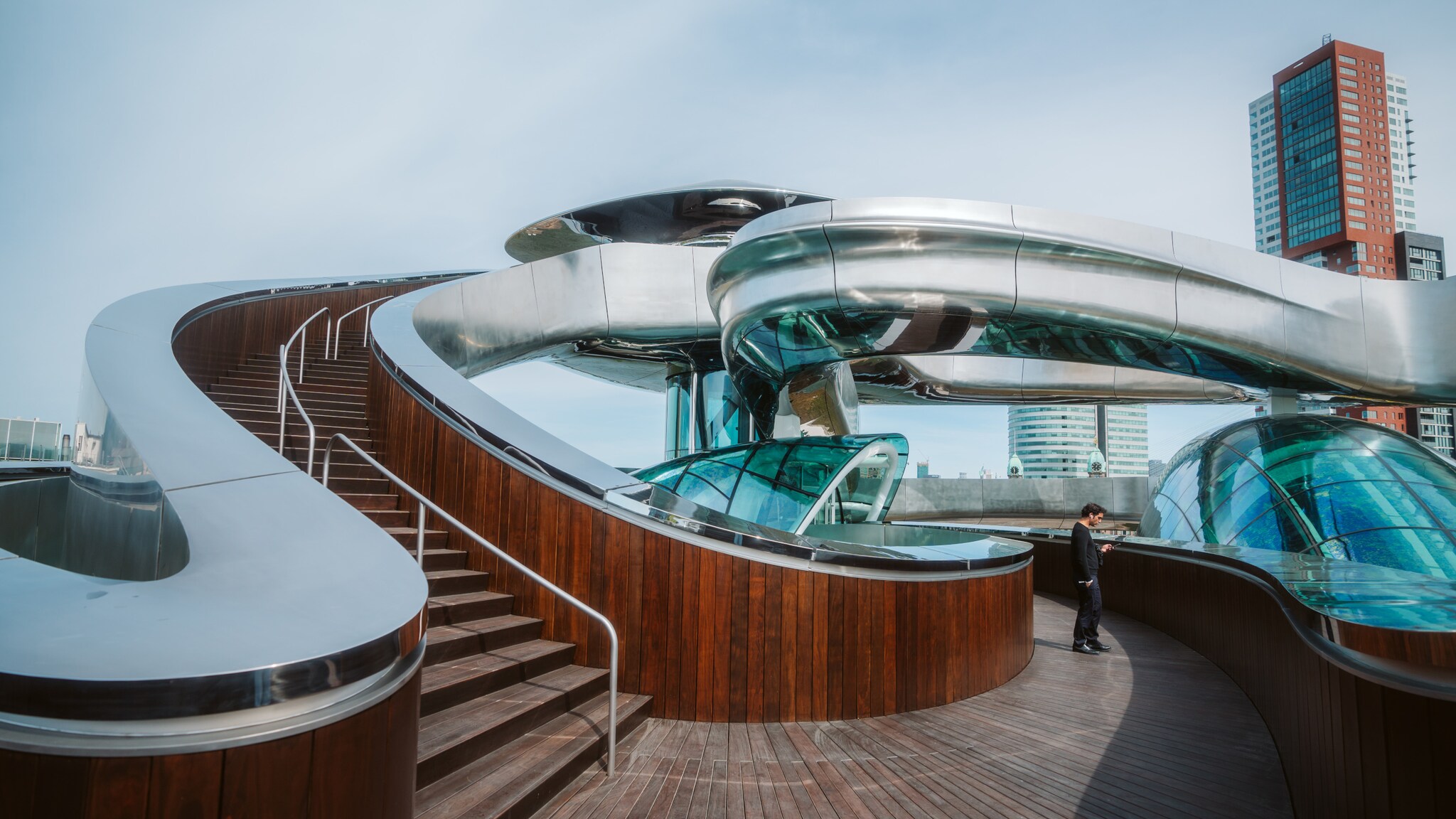 Moderne Dachterrasse mit geschwungenem Holzgeländer, breiter Treppe und silberne, spiralförmige Skulptur “Tornado”, im Hintergrund Hochhäuser.