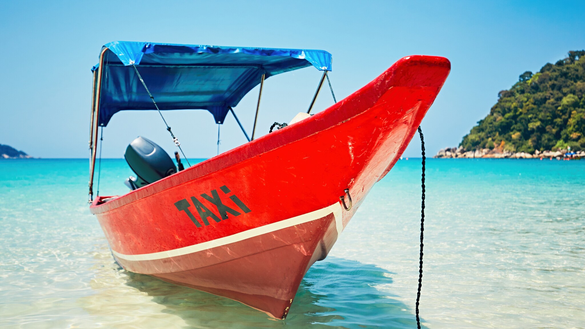 Roter Bootsrumpf mit schwarzer Aufschrift 'TAXI' am Bug, im flachen Wasser vor bewaldetem Ufer und Strand mit Palmen.
