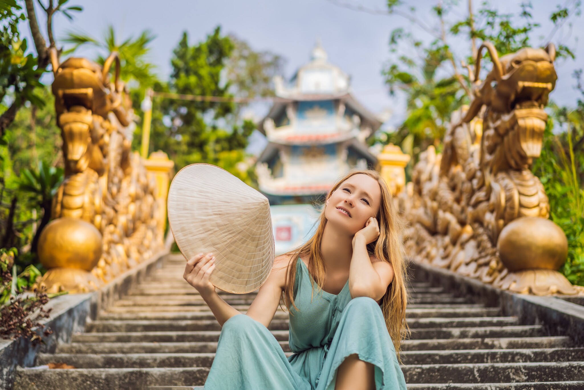 Eine Frau sitzt lächelnd auf einer Treppe. Sie schwenkt einen traditionellen vietnamesischen Hut. Im Hintergrund sind ein Tempel und goldene Statuen zu sehen.