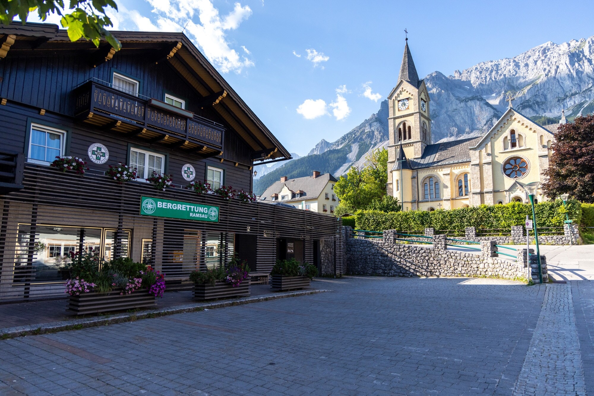 Bergrettungsstation in einem Holzhaus an einem Platz vor einer Kirche in einer Ortschaft mit Bergpanorama im Hintergrund. Bergrettungsstation in einem Holzhaus an einem Platz vor einer Kirche in einer Ortschaft mit Bergpanorama im Hintergrund.