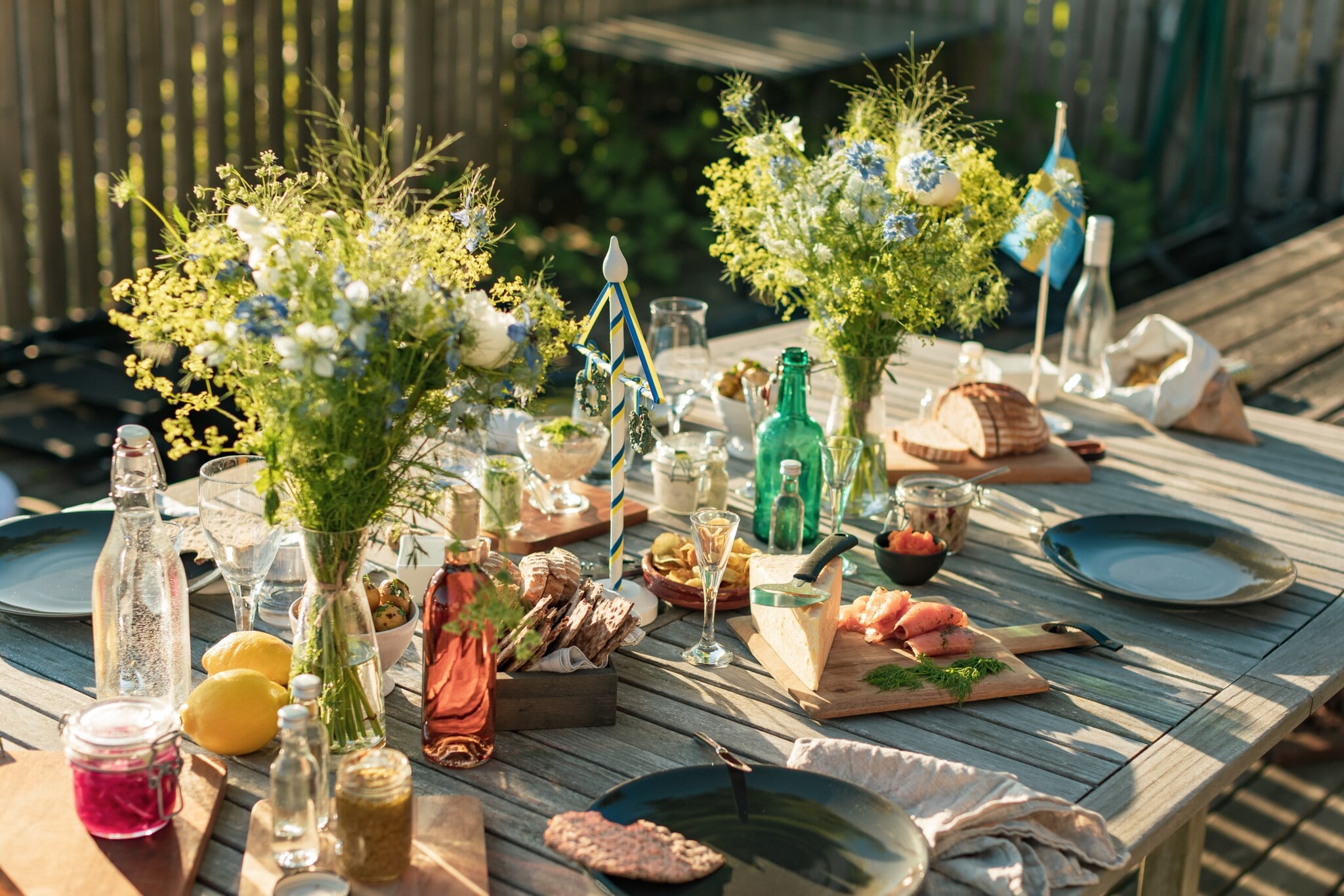 Gedeckter Holztisch für ein Mittsommer-Essen im Freien, mit Brot, gebeiztem Lachs, Käse, Eingelegtem, Getränken und frischen Blumensträußen. Gedeckter Holztisch für ein Mittsommer-Essen im Freien, mit Brot, gebeiztem Lachs, Käse, Eingelegtem, Getränken und frischen Blumensträußen.