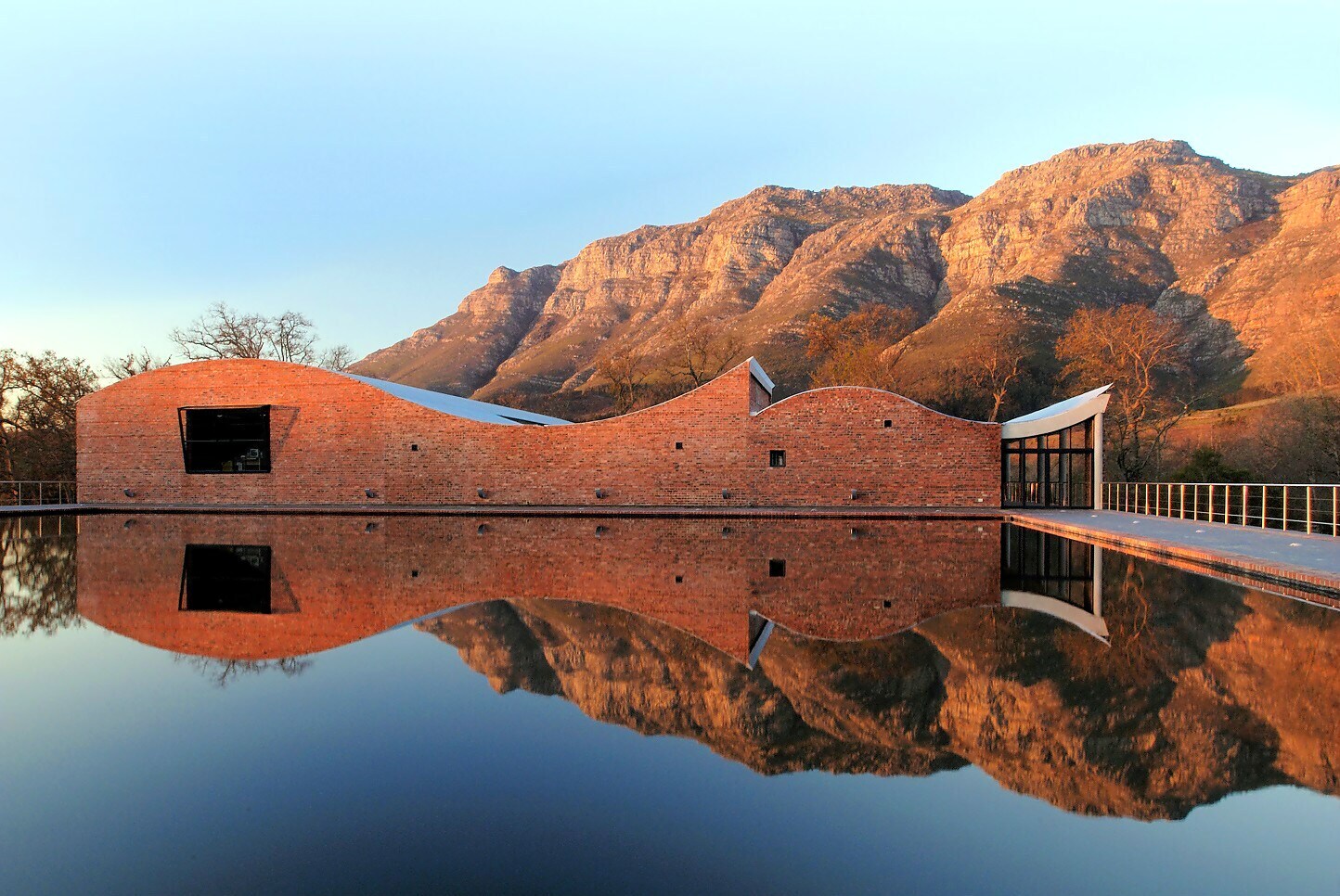 Modernes Gebäude aus rotem Stein mit geschwungenem Dach, das sich in einem Wasserbecken spiegelt, im Hintergrund ein Gebirge in der Abendsonne