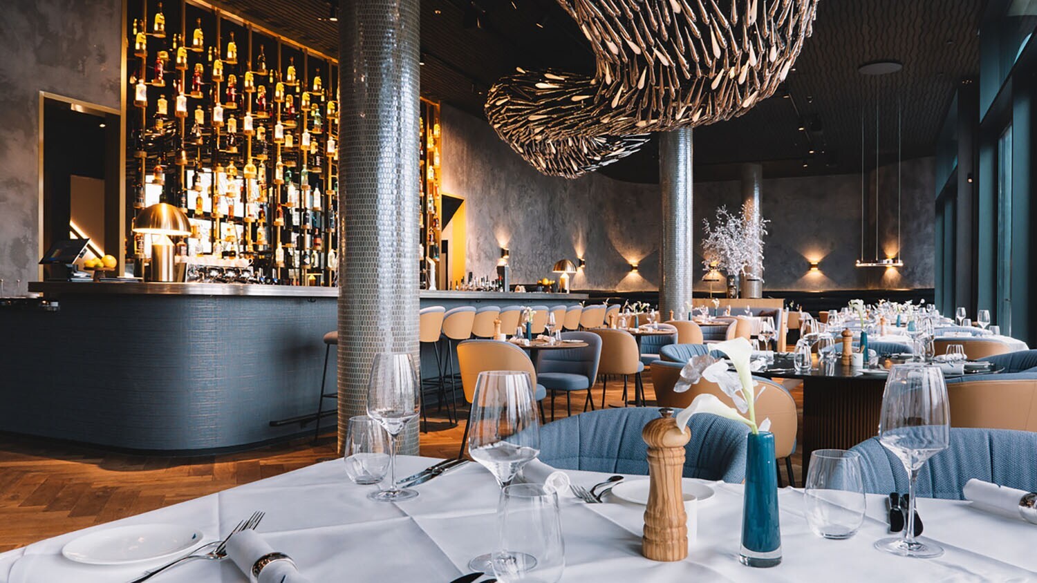 Stilvoller, modern eingerichteter Speisesaal mit Barbereich und eingedeckten Tischen unter einer abstrakten Deckenskulptur.