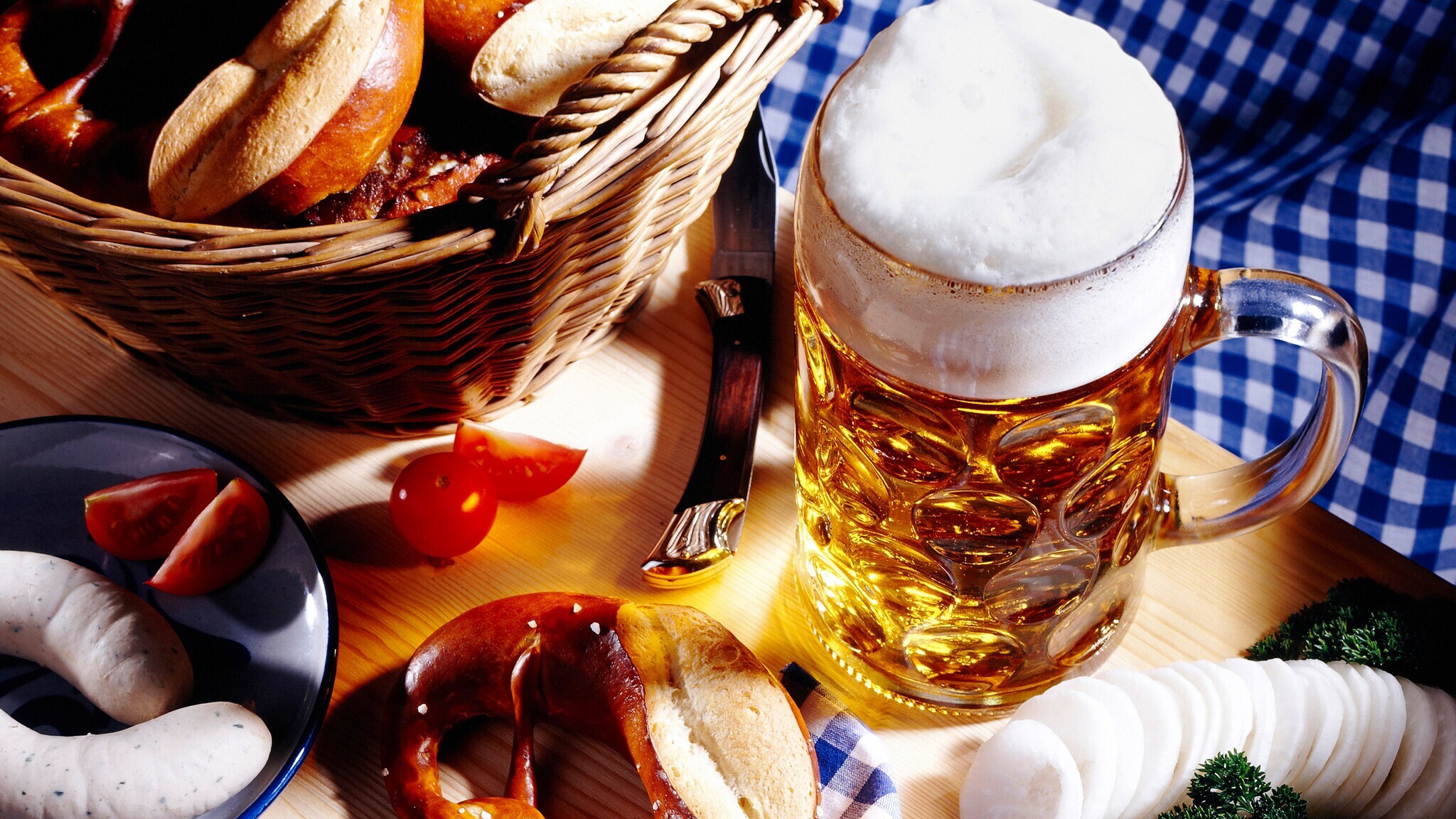 Ein Korb mit Brötchen, ein Glas Bier mit Schaum, eine Brezel, Wurst, Tomaten, Senf und Käse auf einem Holztisch mit kariertem Tuch.