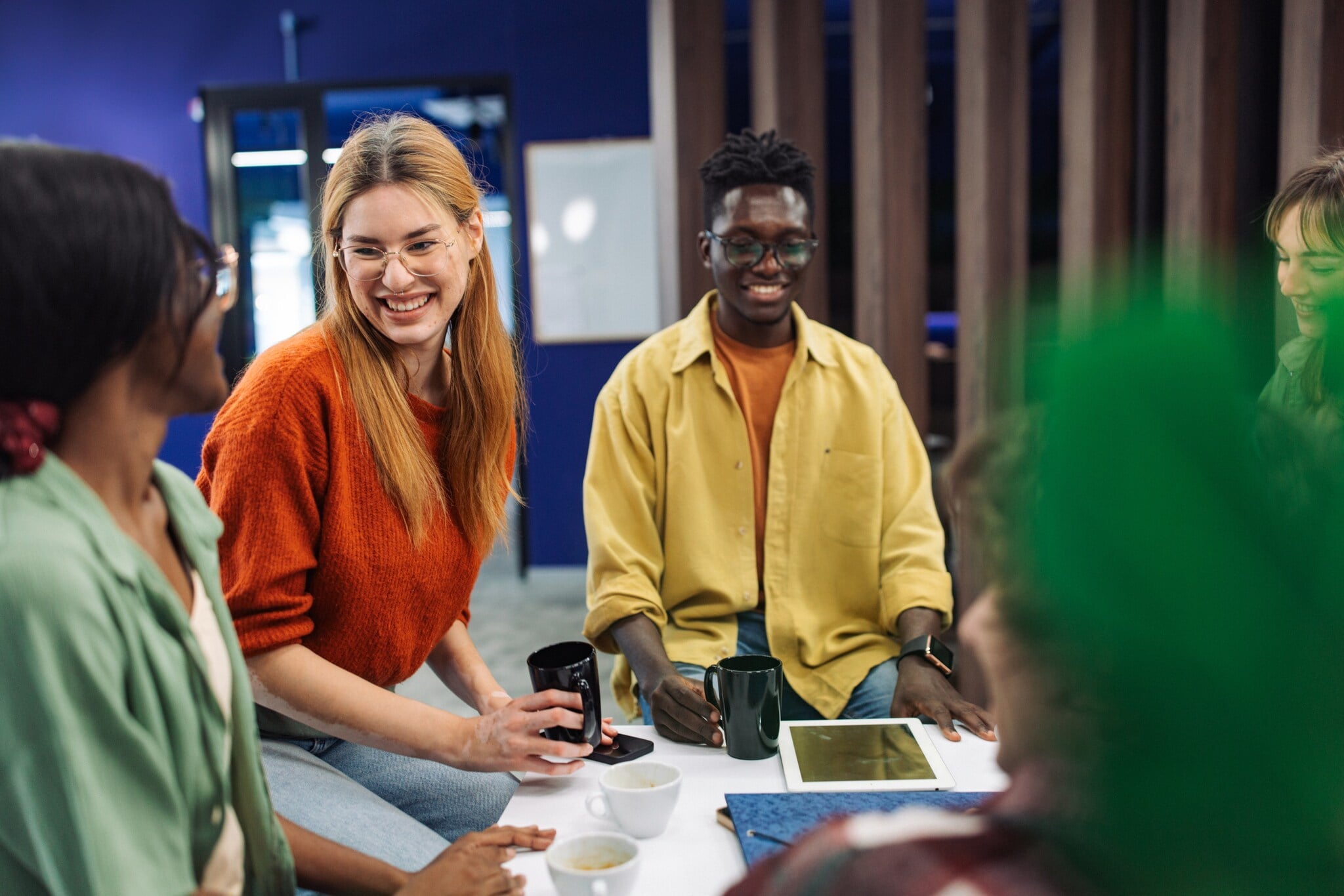 Eine Gruppe junger Menschen sitzt lächelnd beieinander, zwischen ihnen Kaffeetassen und Tablets auf einem Tisch. Eine Gruppe junger Menschen sitzt lächelnd beieinander, zwischen ihnen Kaffeetassen und Tablets auf einem Tisch.