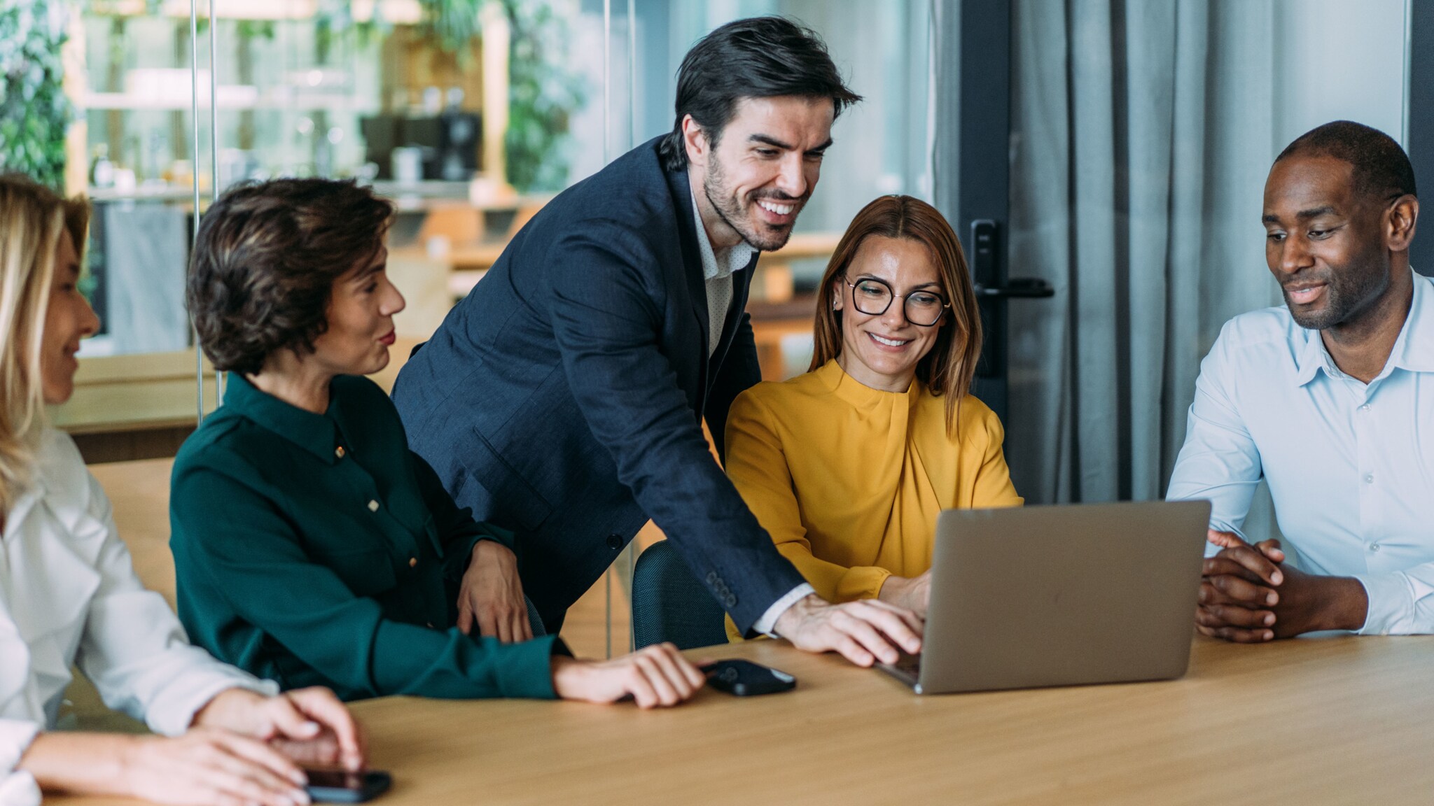 Ein Mann im Anzug bedient stehend einen Laptop an einem Konferenztisch mit vier sitzenden Personen. Ein Mann im Anzug bedient stehend einen Laptop an einem Konferenztisch mit vier sitzenden Personen.