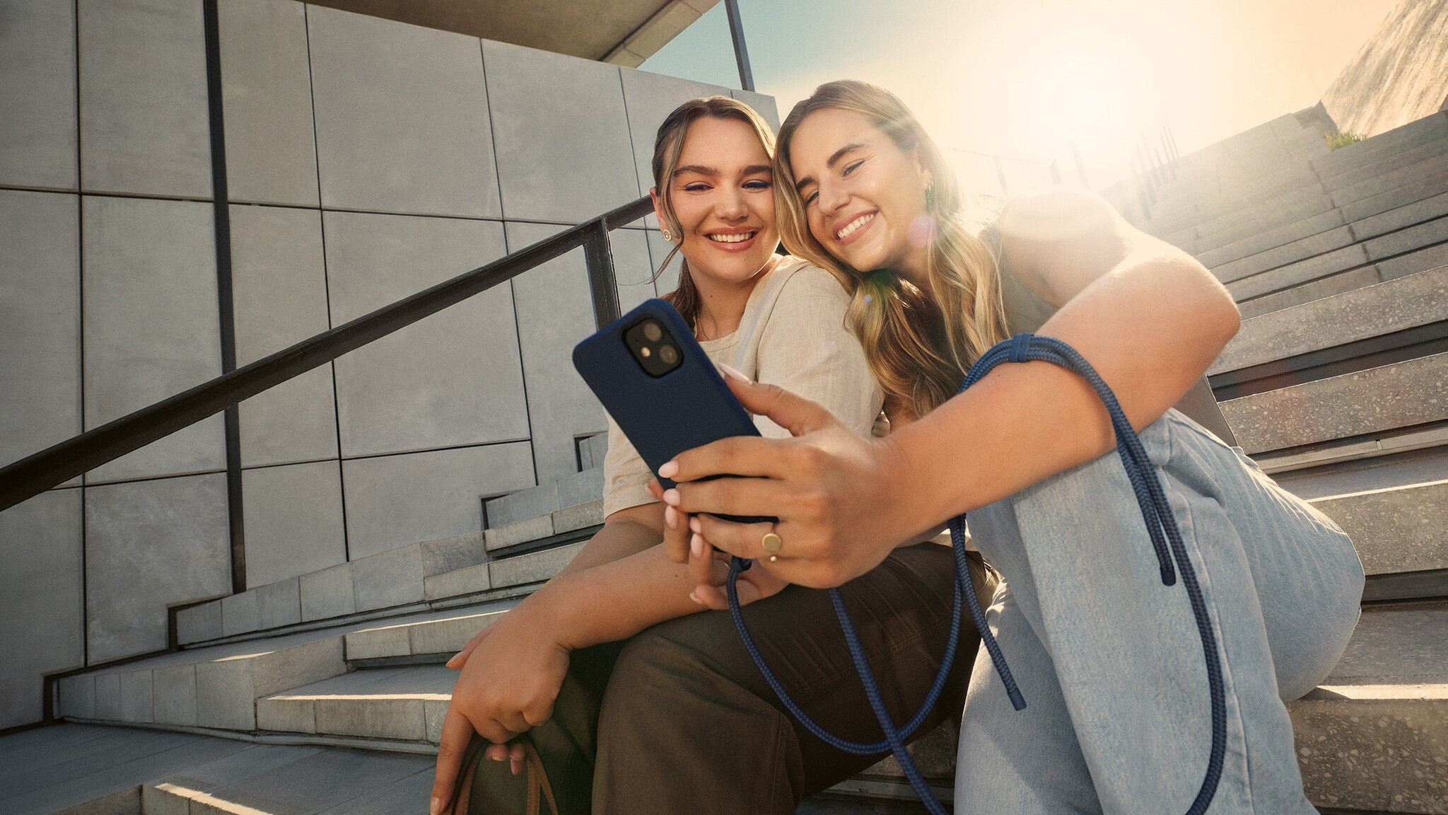 Zwei junge Frauen sitzen auf einer großen Treppe und schauen lächelnd auf ein Smartphone