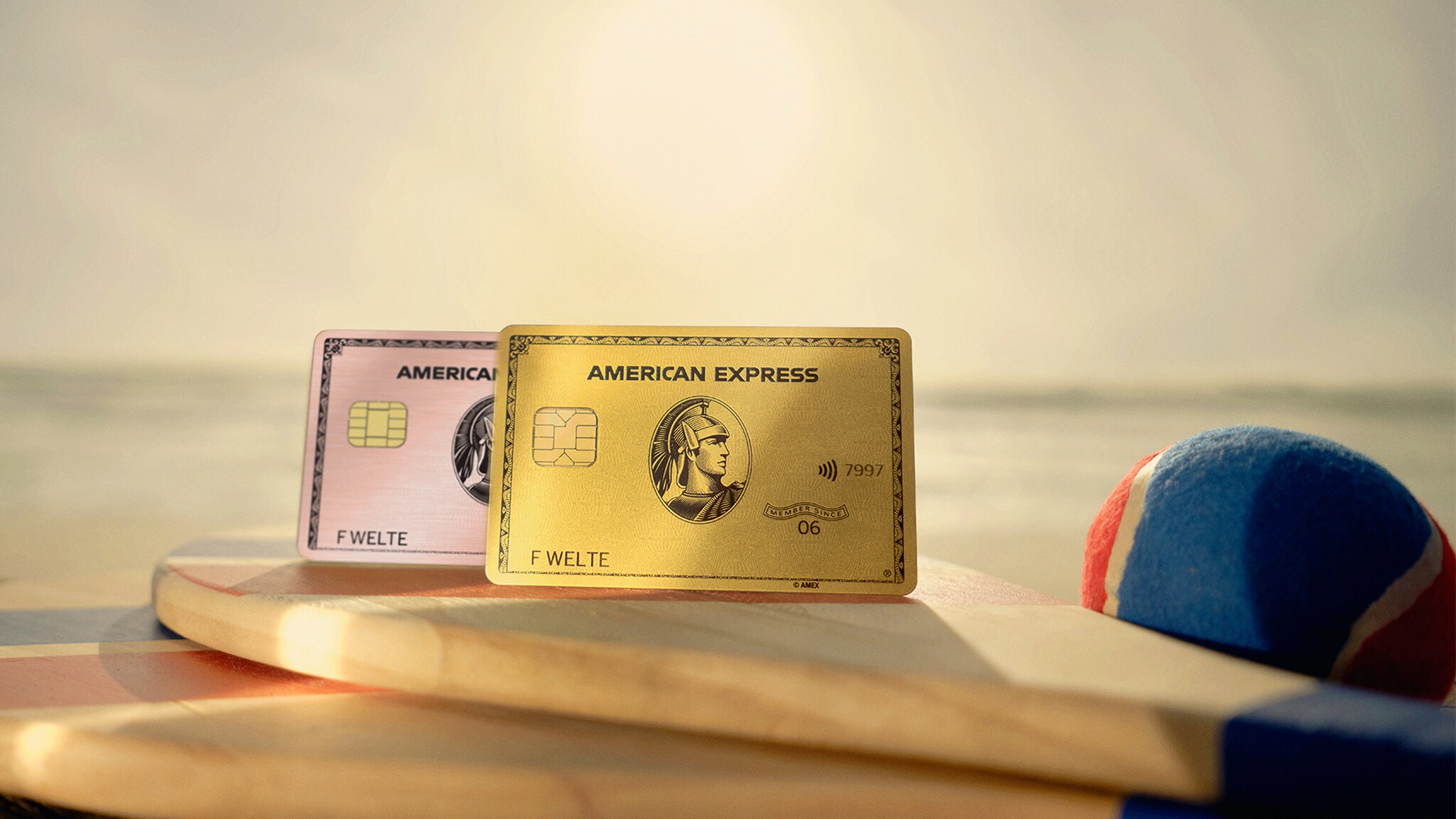 Zwei Kreditkarten von American Express liegen auf einem Holzschläger am Strand, im Hintergrund sieht man das Meer bei Sonnenuntergang.