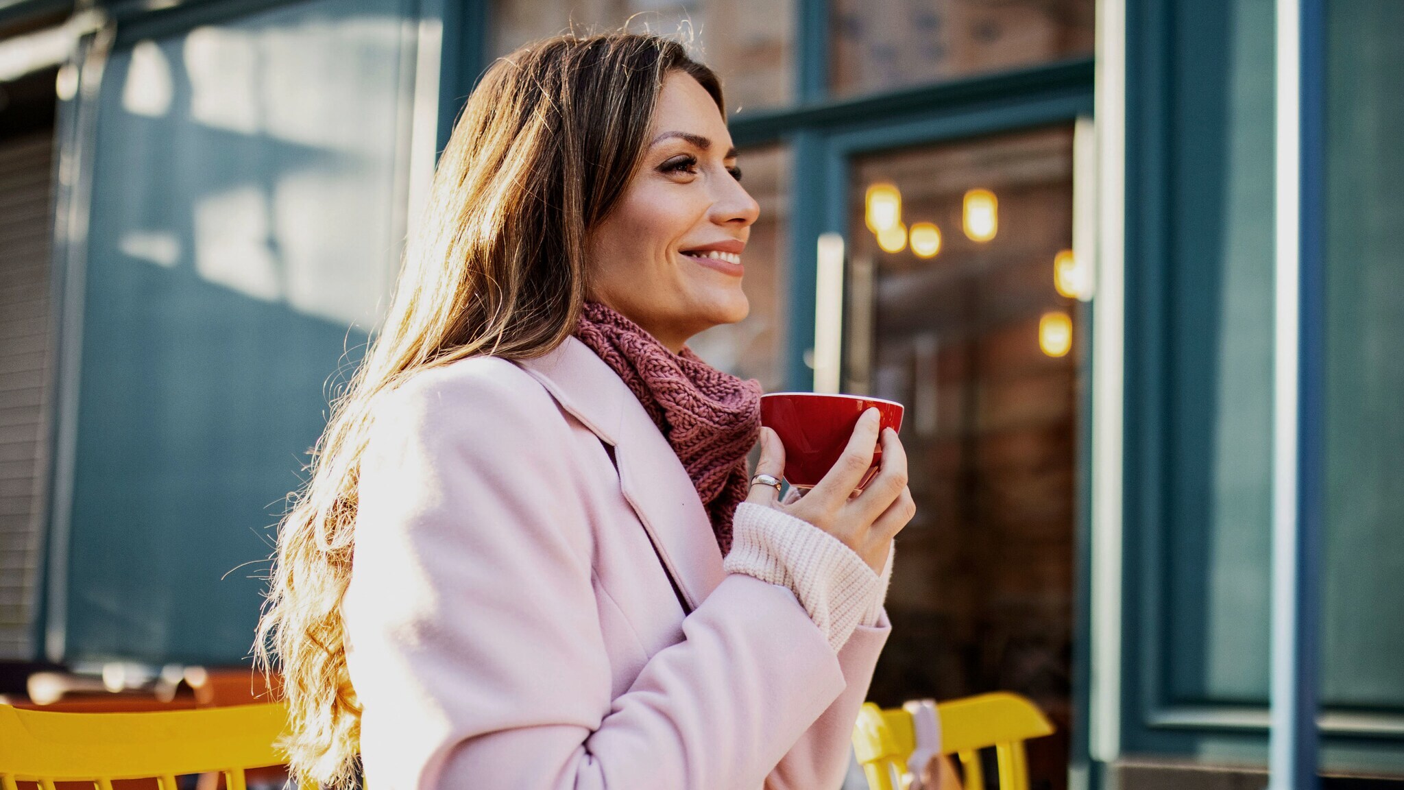 Eine Frau im rosafarbenen Mantel sitzt zufrieden lächelnd mit einer roten Cappuccinotasse in den Händen auf der Außenterrasse eines Cafés im Sonnenschein.