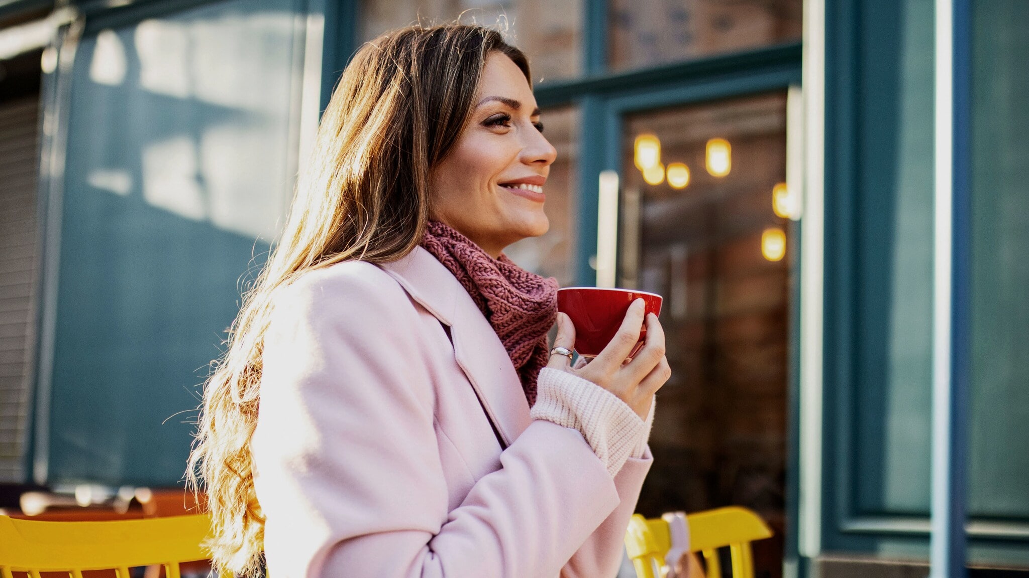 Eine Frau im rosafarbenen Mantel sitzt zufrieden lächelnd mit einer roten Cappuccinotasse in den Händen auf der Außenterrasse eines Cafés im Sonnenschein. Eine Frau im rosafarbenen Mantel sitzt zufrieden lächelnd mit einer roten Cappuccinotasse in den Händen auf der Außenterrasse eines Cafés im Sonnenschein.