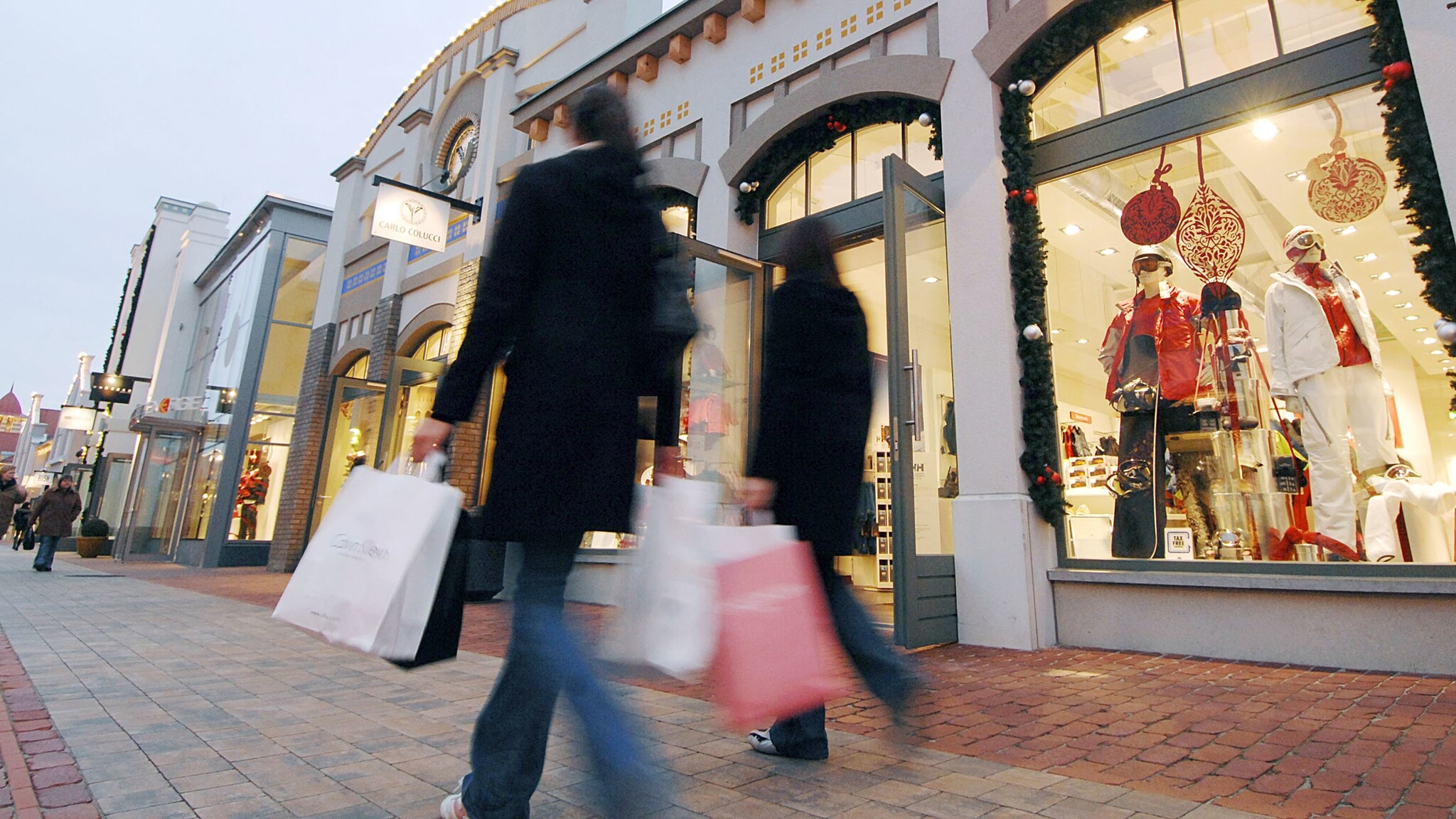 Zwei Personen in Mänteln mit Einkaufstaschen laufen vor beleuchteten Schaufenstern von Modegeschäften in einer Einkaufsstraße vorbei. Zwei Personen in Mänteln mit Einkaufstaschen laufen vor beleuchteten Schaufenstern von Modegeschäften in einer Einkaufsstraße vorbei.