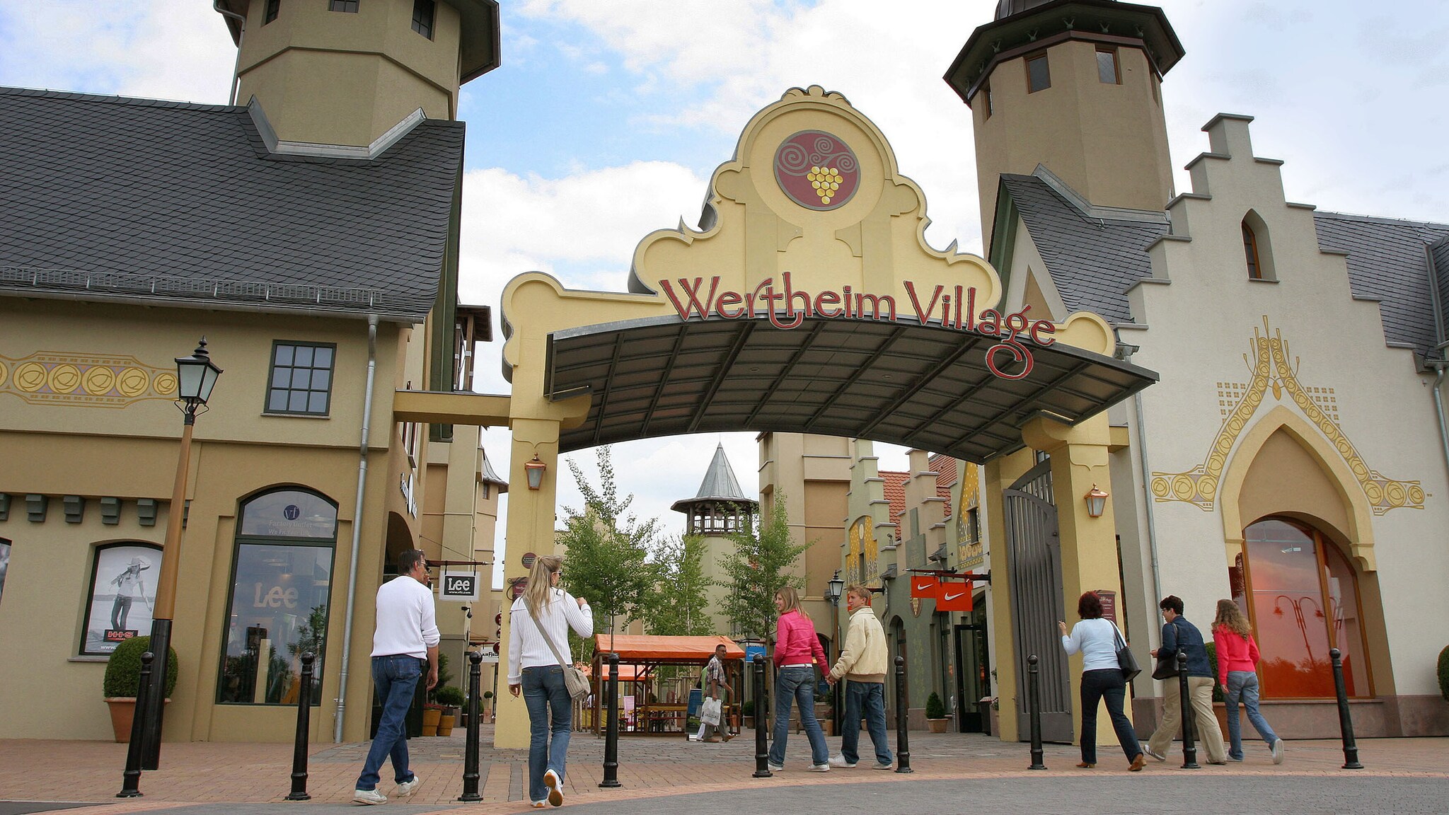 Personen an einem Eingangsbereich mit der Aufschrift „Wertheim Village“ auf einem Torbogen zwischen zwei Türmen eines Einkaufszentrums unter freiem Himmel.