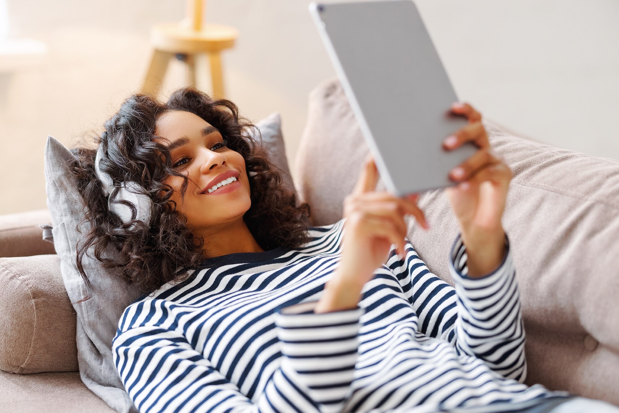 Delighted ethnic female smiling and browsing tablet while resting on couch and listening to music in headphones at home