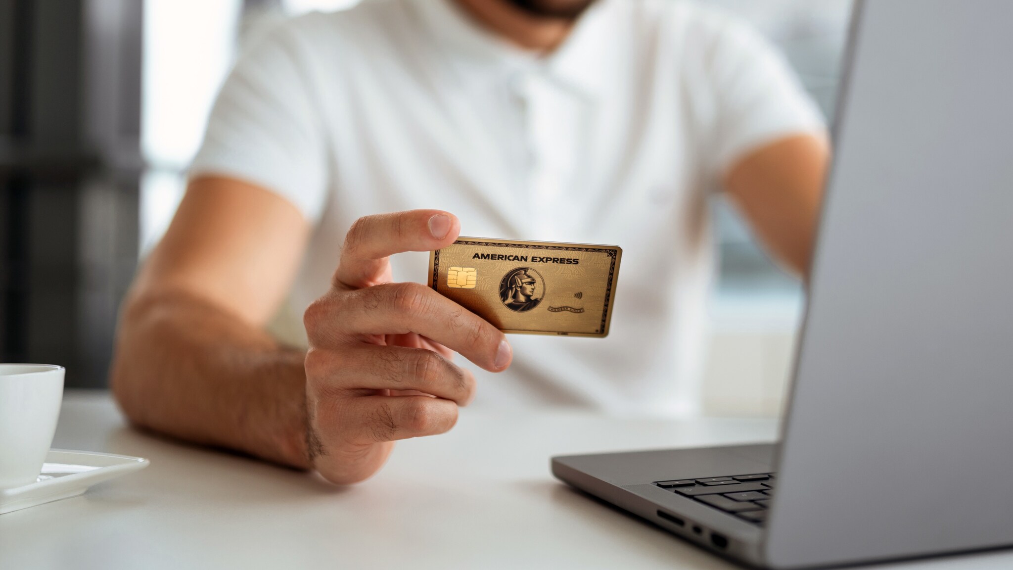 Nahaufnahme einer goldenen American Express Kreditkarte in der Hand einer Person bei der Online-Zahlung an einem Laptop.