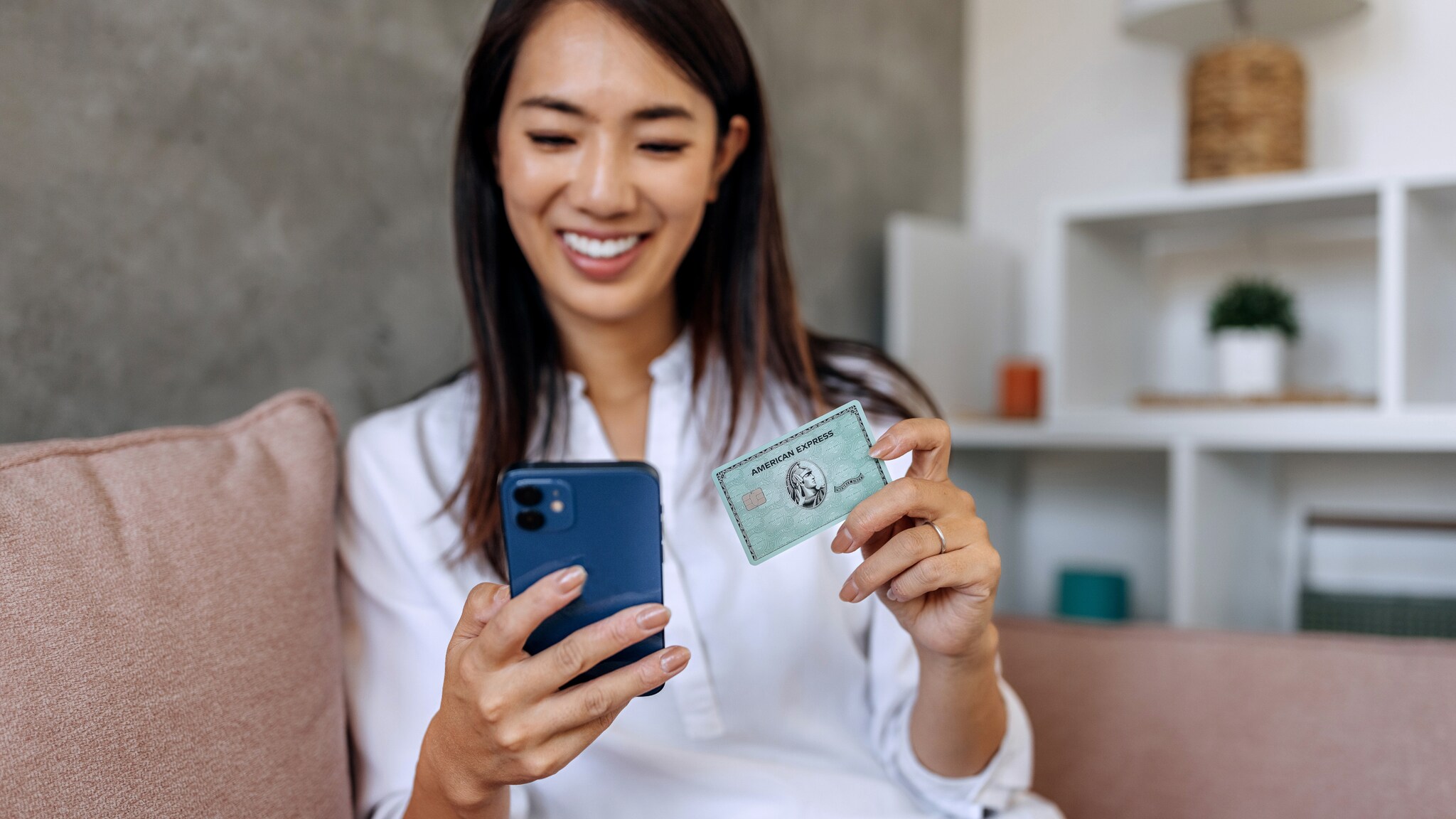 Eine Frau sitzt auf einem Sofa und blickt lächelnd auf ein Smartphone in ihrer Hand, in der anderen Hand hält sie eine grüne American Express Kreditkarte.