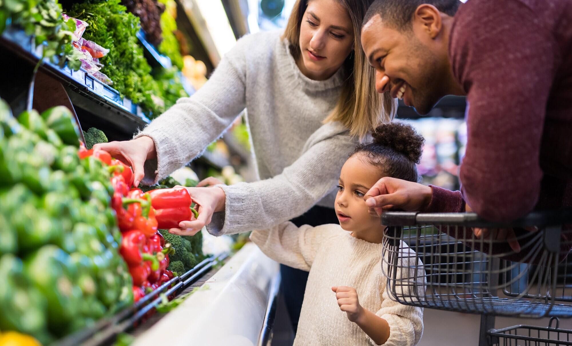 Ein kleines Mädchen steht mit seinen Eltern im Supermarkt vor dem Gemüseregal und sucht Paprika aus