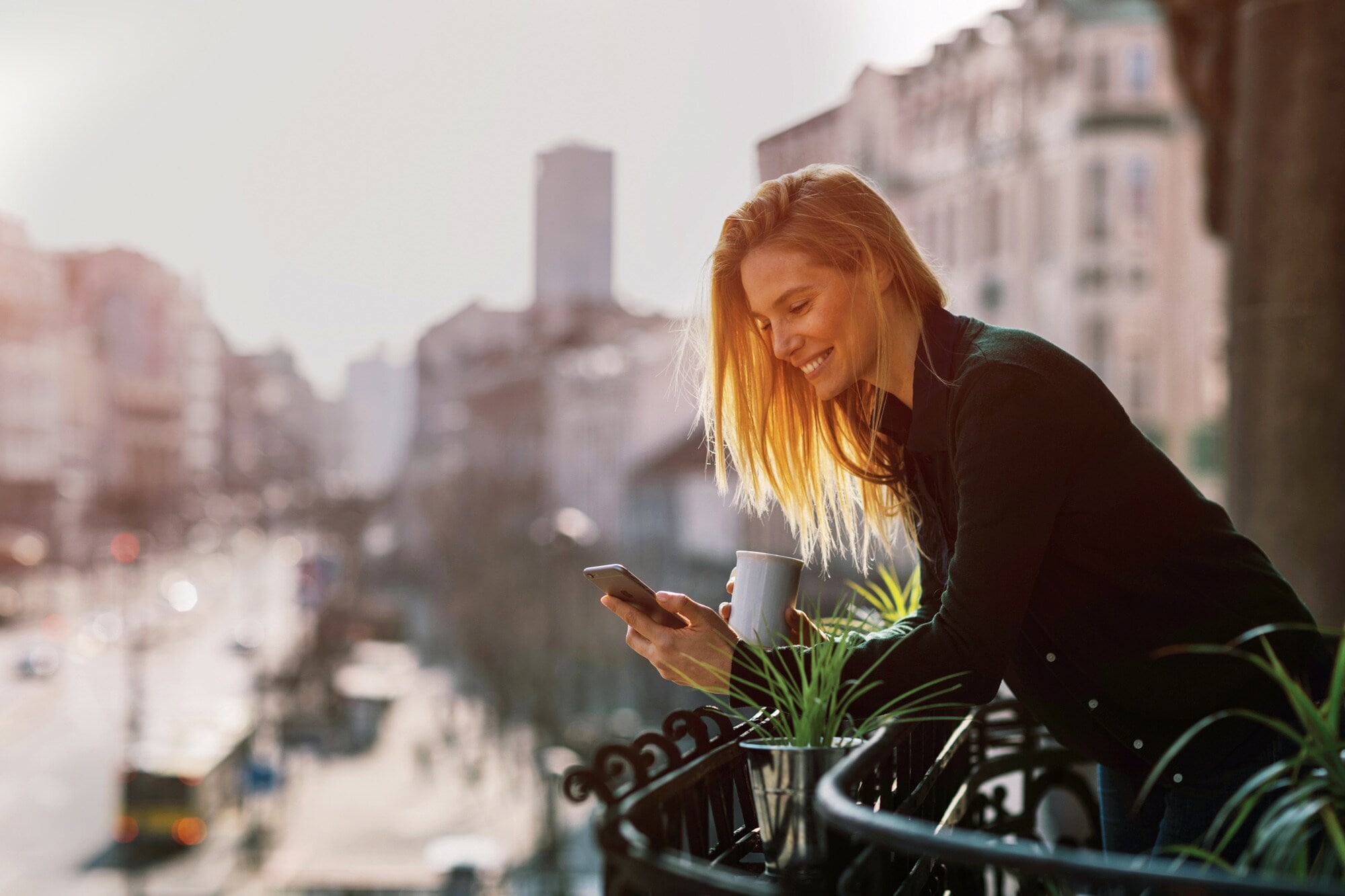 Eine Frau steht mit einer Kaffeetasse auf einem Balkon und schaut auf ihr Smartphone Eine Frau steht mit einer Kaffeetasse auf einem Balkon und schaut auf ihr Smartphone
