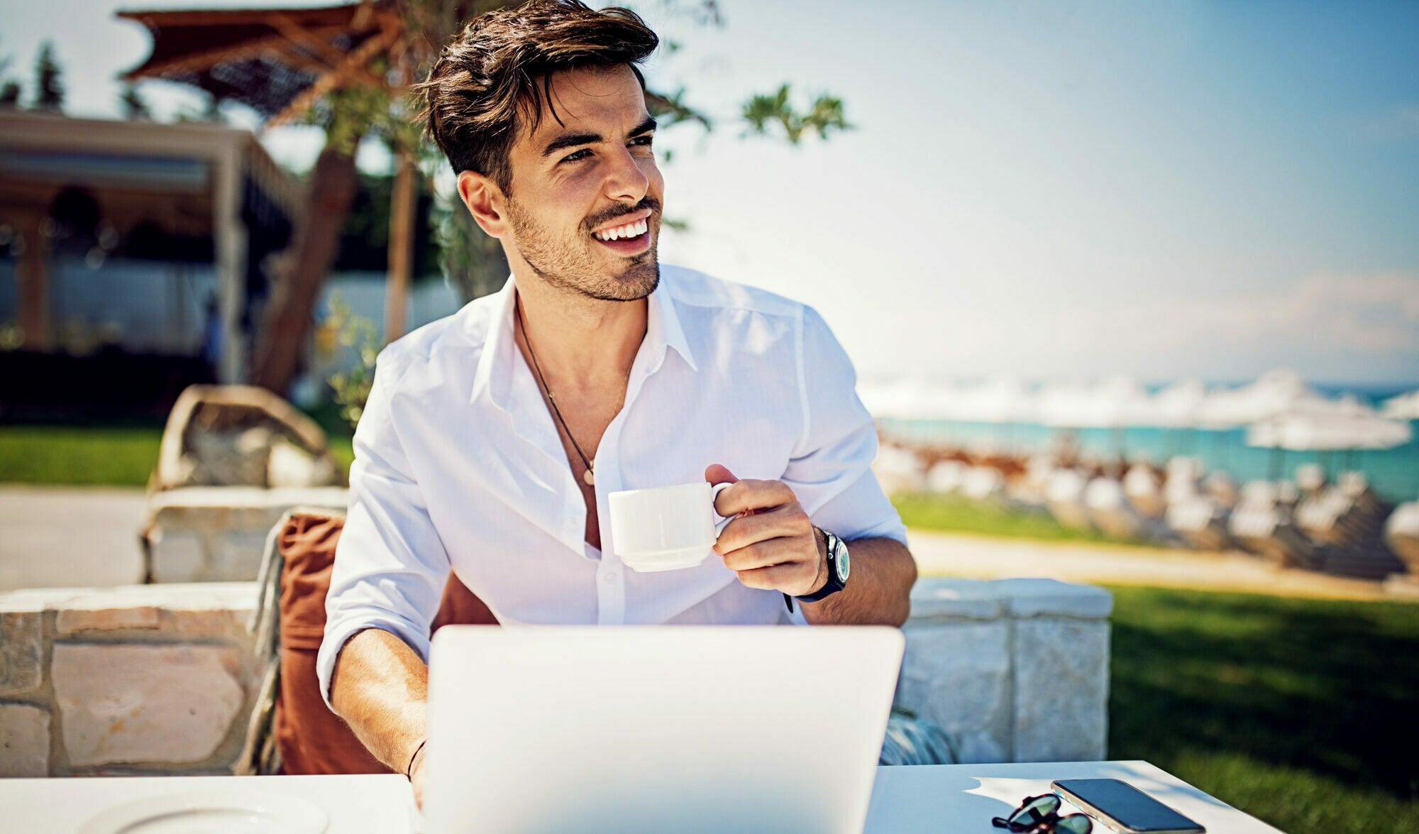 Ein junger Mann im weißen Hemd sitzt an einem Strand mit einer Kaffeetasse vor einem Laptop in der Sonne Ein junger Mann im weißen Hemd sitzt an einem Strand mit einer Kaffeetasse vor einem Laptop in der Sonne