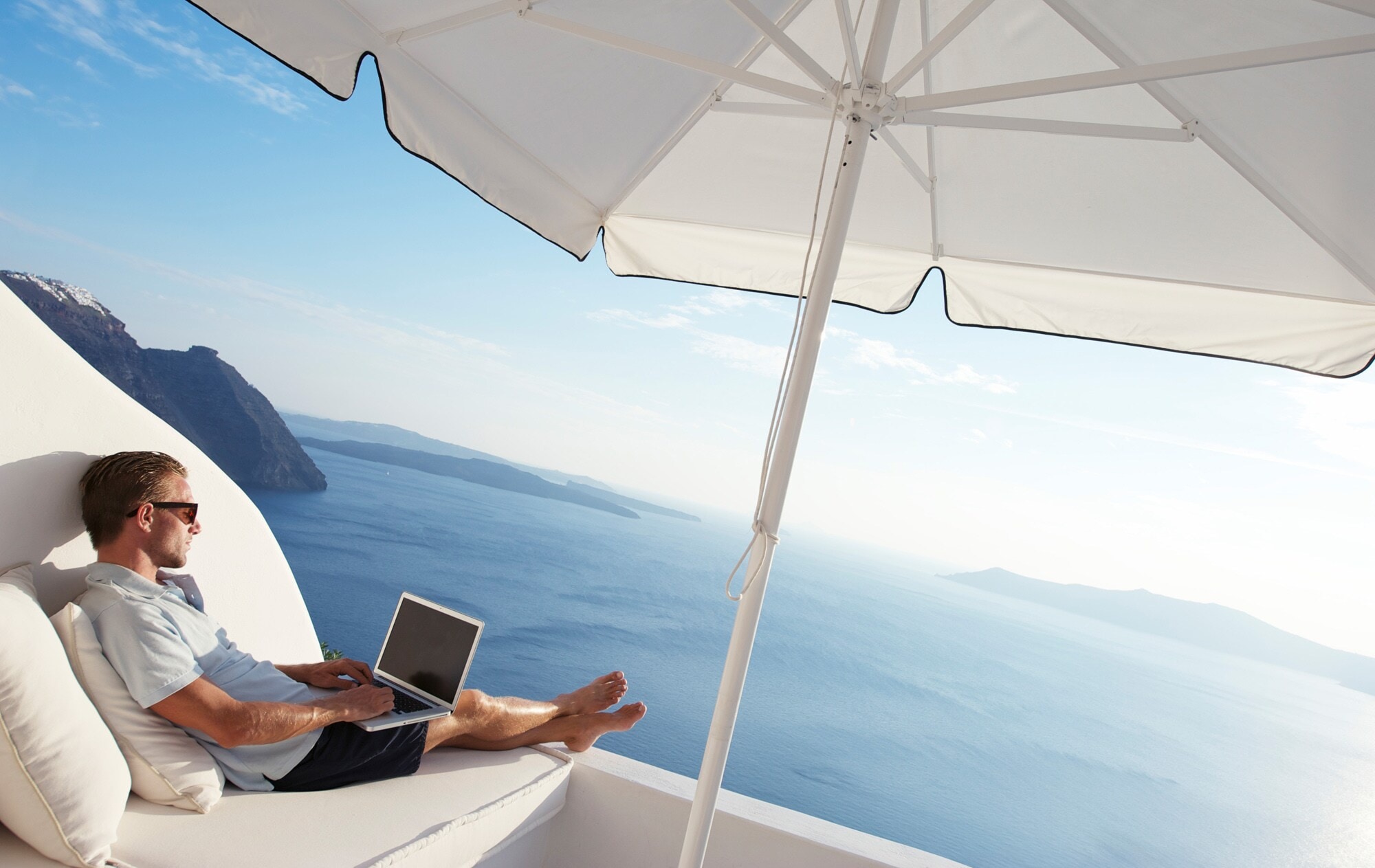 Ein leger gekleideter Mann mit Laptop auf dem Schoß auf einer luxuriösen Terrasse mit Meerblick Ein leger gekleideter Mann mit Laptop auf dem Schoß auf einer luxuriösen Terrasse mit Meerblick