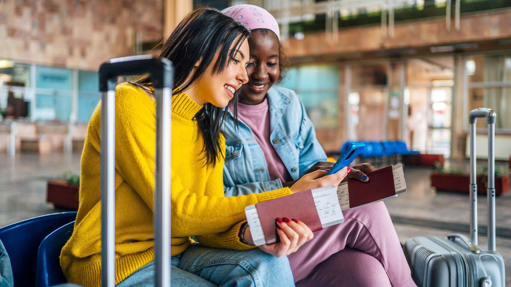 Zwei junge Frauen sitzen mit Trolleys, Reisepässen und Bordkarten in einem Terminal und schauen gemeinsam auf ein Smartphone. Zwei junge Frauen sitzen mit Trolleys, Reisepässen und Bordkarten in einem Terminal und schauen gemeinsam auf ein Smartphone.