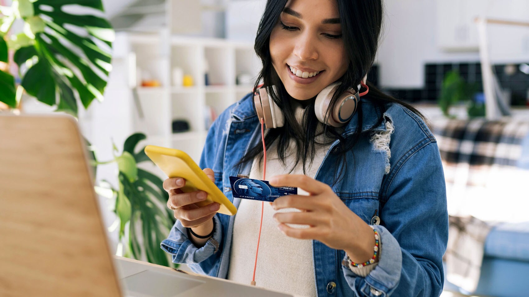 Eine junge Frau mit Kopfhörern sitzt mit einem Smartphone und einer blauen American Express Kreditkarte in den Händen vor einem Laptop am Schreibtisch in einer Wohnung. Eine junge Frau mit Kopfhörern sitzt mit einem Smartphone und einer blauen American Express Kreditkarte in den Händen vor einem Laptop am Schreibtisch in einer Wohnung.