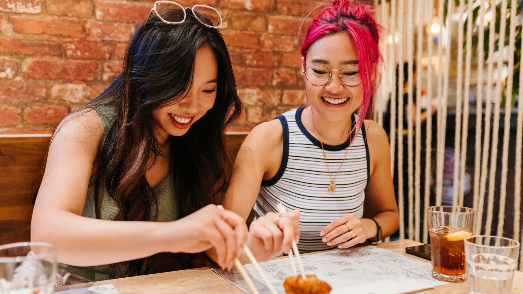 Zwei junge asiatische Frauen an einem Tisch in einem Restaurant greifen mit Essstäbchen nach einer Speise. Zwei junge asiatische Frauen an einem Tisch in einem Restaurant greifen mit Essstäbchen nach einer Speise.