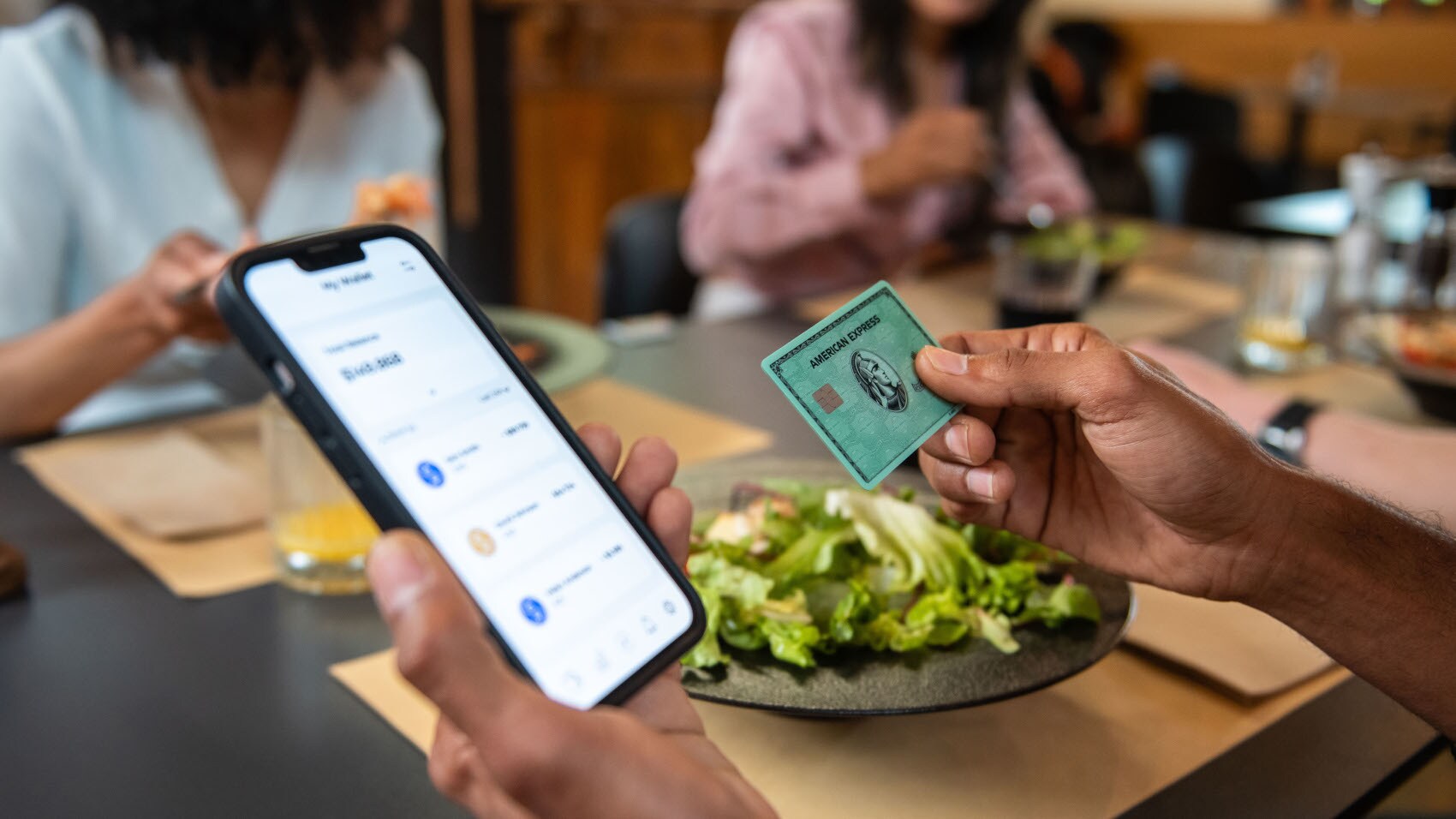 Eine grüne American Express Kreditkarte und ein Smartphone mit Amex App auf dem Display in den Händen einer Person an einem Tisch im Restaurant. Eine grüne American Express Kreditkarte und ein Smartphone mit Amex App auf dem Display in den Händen einer Person an einem Tisch im Restaurant.