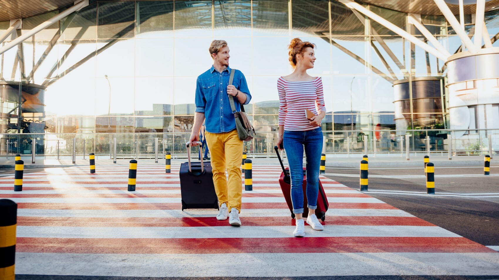 Ein Mann und eine Frau laufen mit Rollkoffern über einen Zebrastreifen vor einem Flughafenterminal.