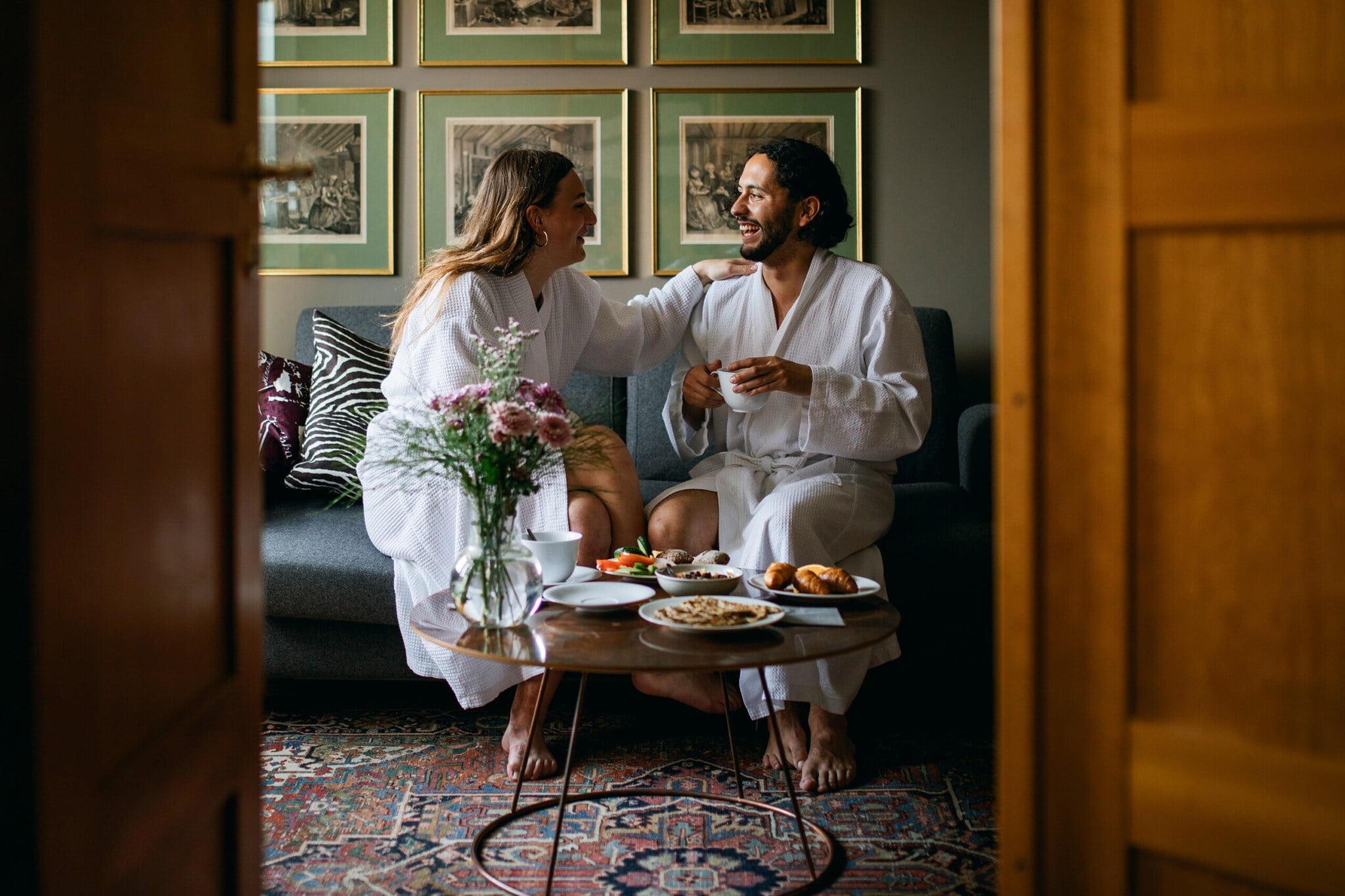 Ein Paar sitzt gut gelaunt in weißen Bademänteln auf einem Sofa in einer stylishen Suite eines Boutiquehotels und genießt das gemeinsame Frühstück Ein Paar sitzt gut gelaunt in weißen Bademänteln auf einem Sofa in einer stylishen Suite eines Boutiquehotels und genießt das gemeinsame Frühstück