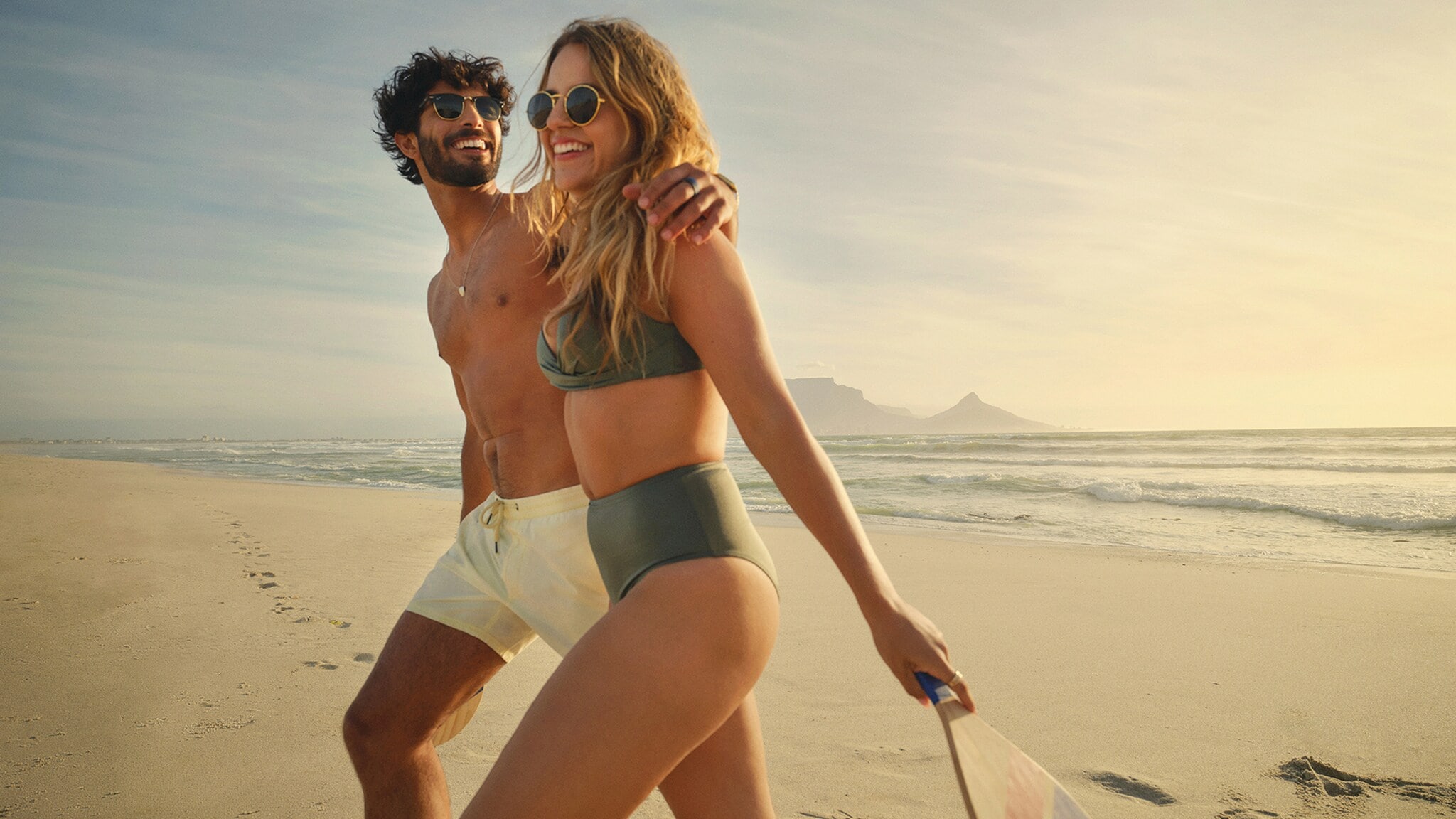 Ein Paar läuft lächelnd Arm in Arm am weitläufigen Strand entlang, in der Hand hält die Frau einen Beach-Schläger aus Holz. Ein Paar läuft lächelnd Arm in Arm am weitläufigen Strand entlang, in der Hand hält die Frau einen Beach-Schläger aus Holz.