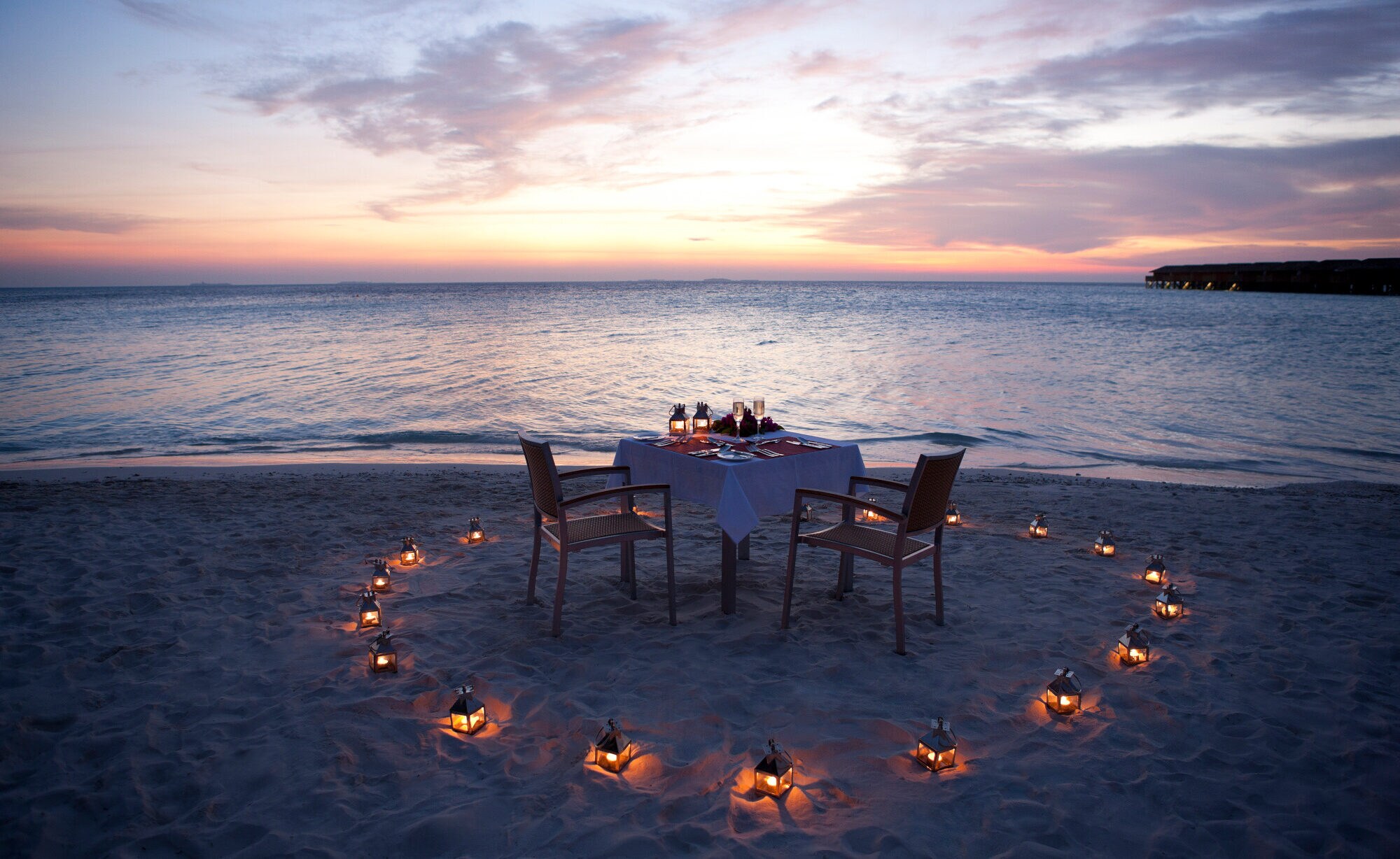 Ein gedeckter Tisch am Strand eines Luxusresorts, umgeben von Kerzen bei Sonnenuntergang Ein gedeckter Tisch am Strand eines Luxusresorts, umgeben von Kerzen bei Sonnenuntergang